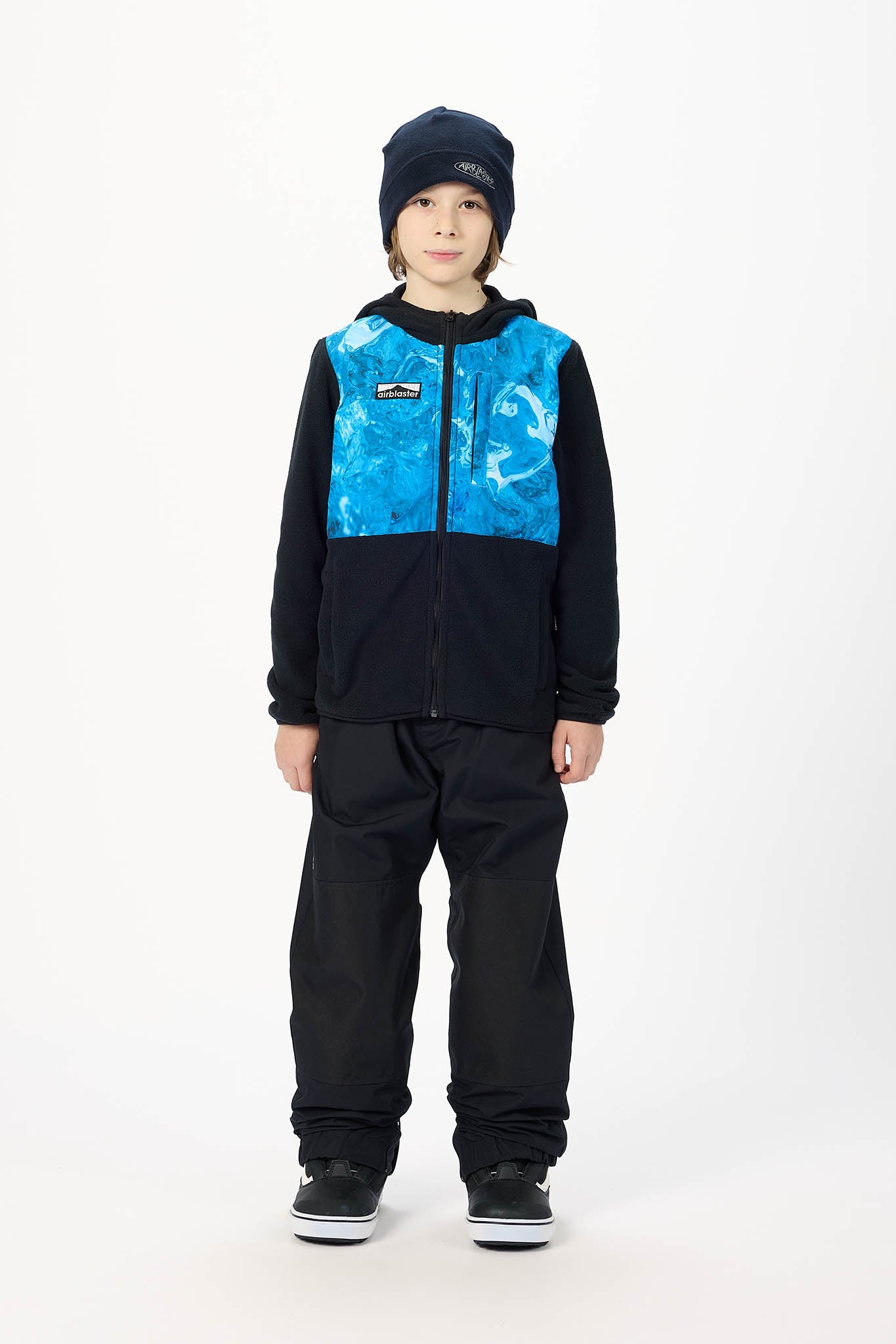 A child model wearing a Patagonia kids' zip-up fleece jacket in black and blue, paired with black pants, a black beanie, and black sneakers, standing against a white studio background.