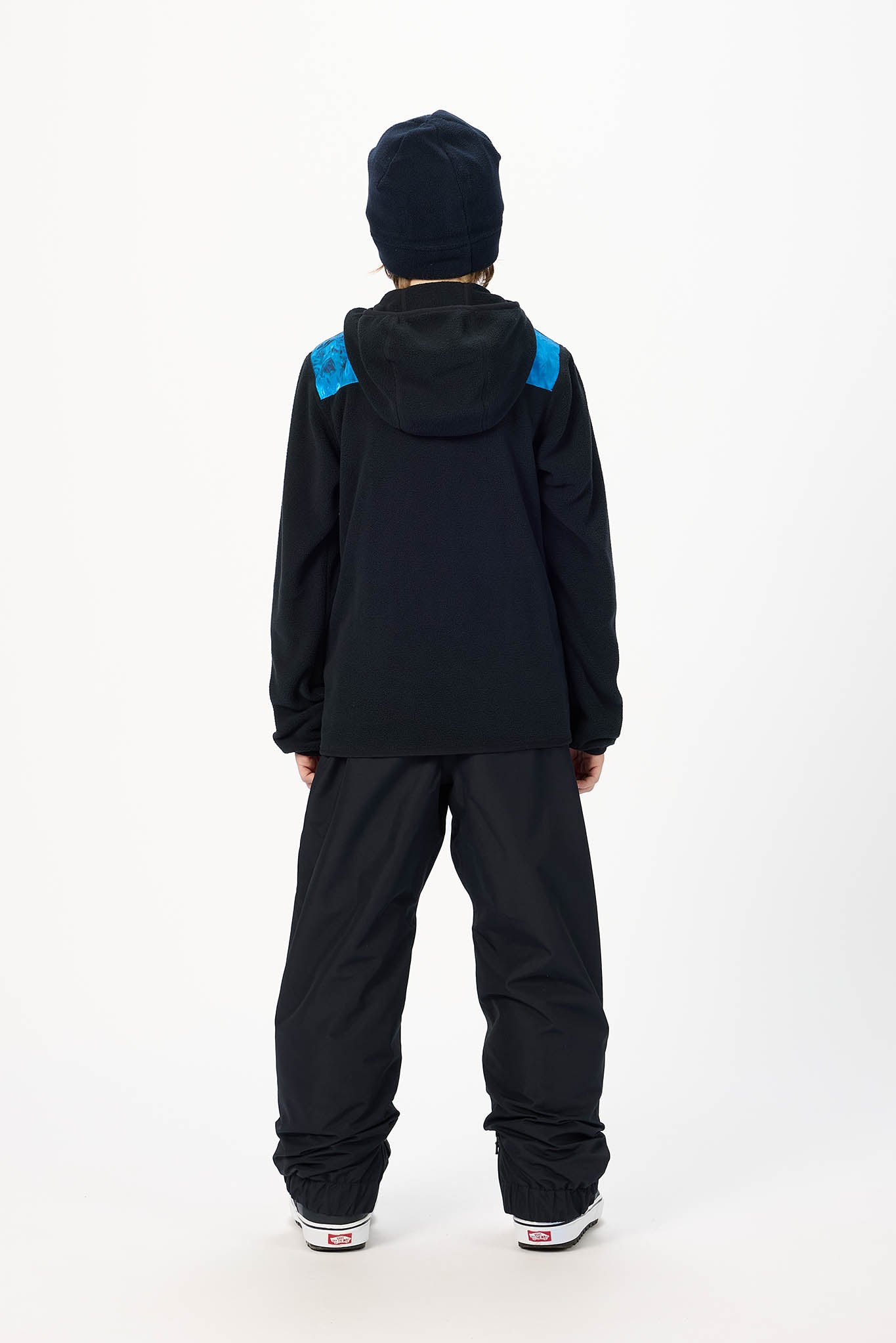 A child shown from the back wearing a black hooded jacket with bright blue shoulder panels, black snow pants, and a black beanie against a plain white studio background.