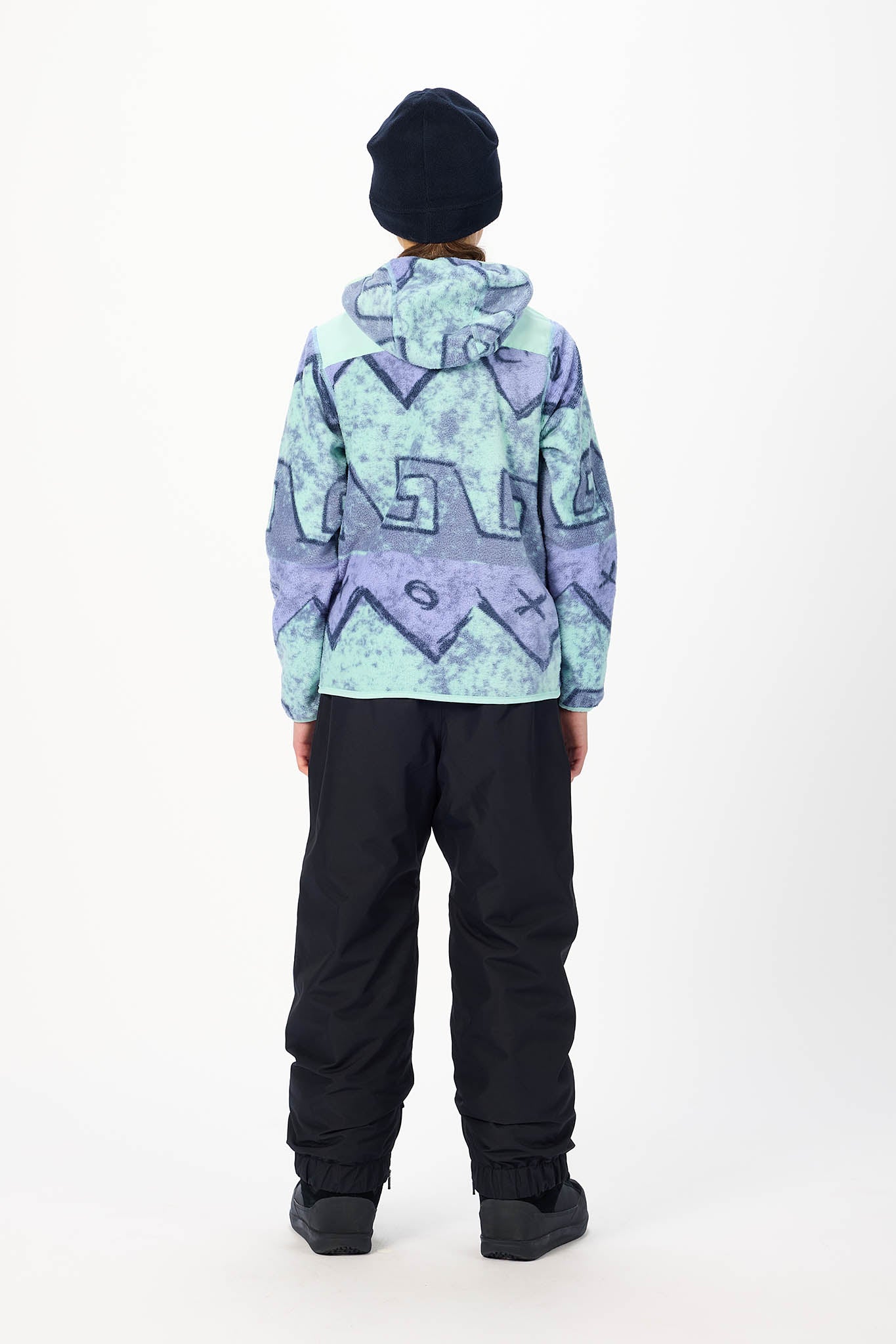 A child viewed from behind wearing a hooded jacket with a light blue and purple geometric pattern, black pants, and a black beanie against a plain white studio background.