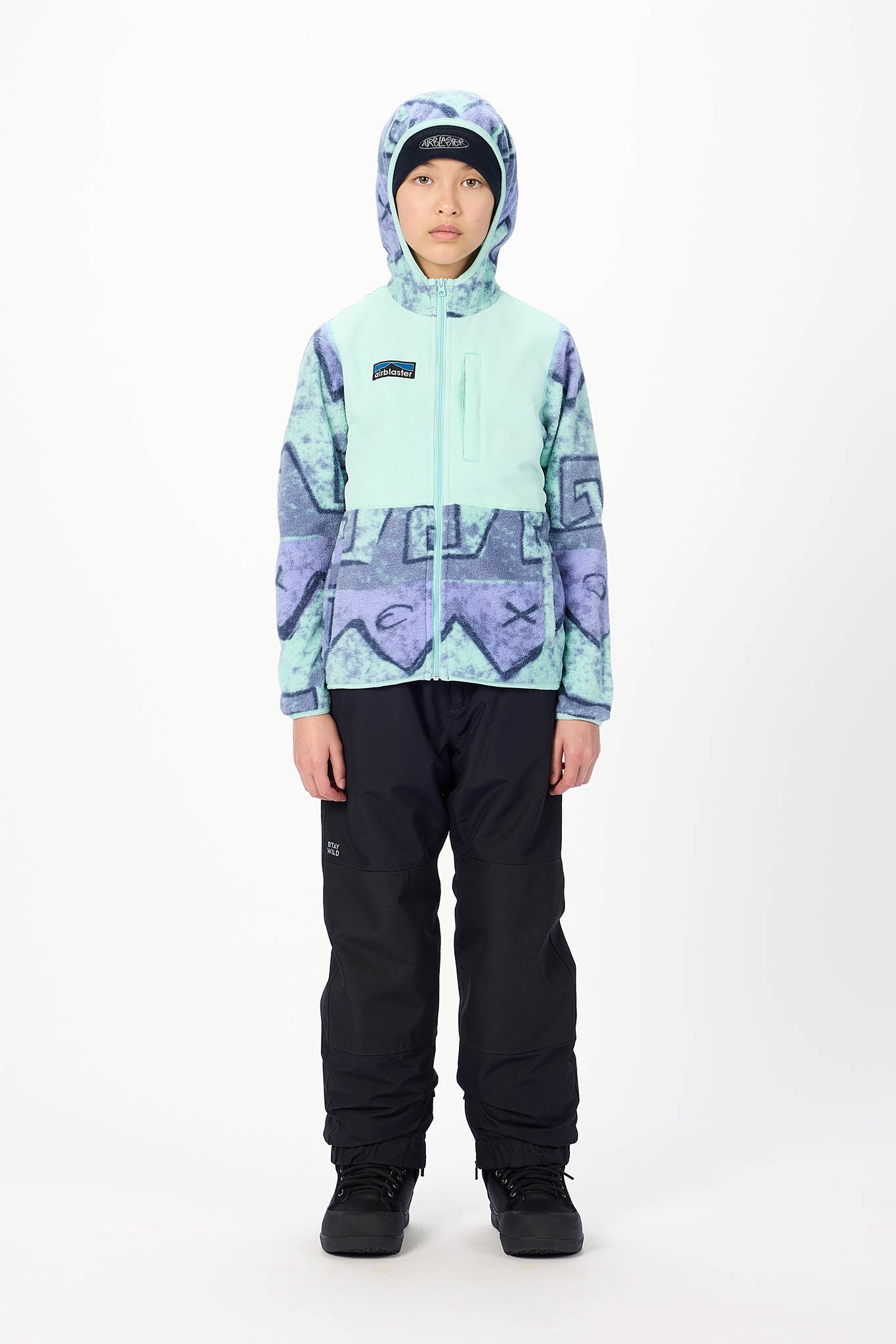 A child model wearing a Patagonia kids' hooded snow jacket in a light mint green and purple geometric pattern, paired with black snow pants and black boots, standing against a white studio background.