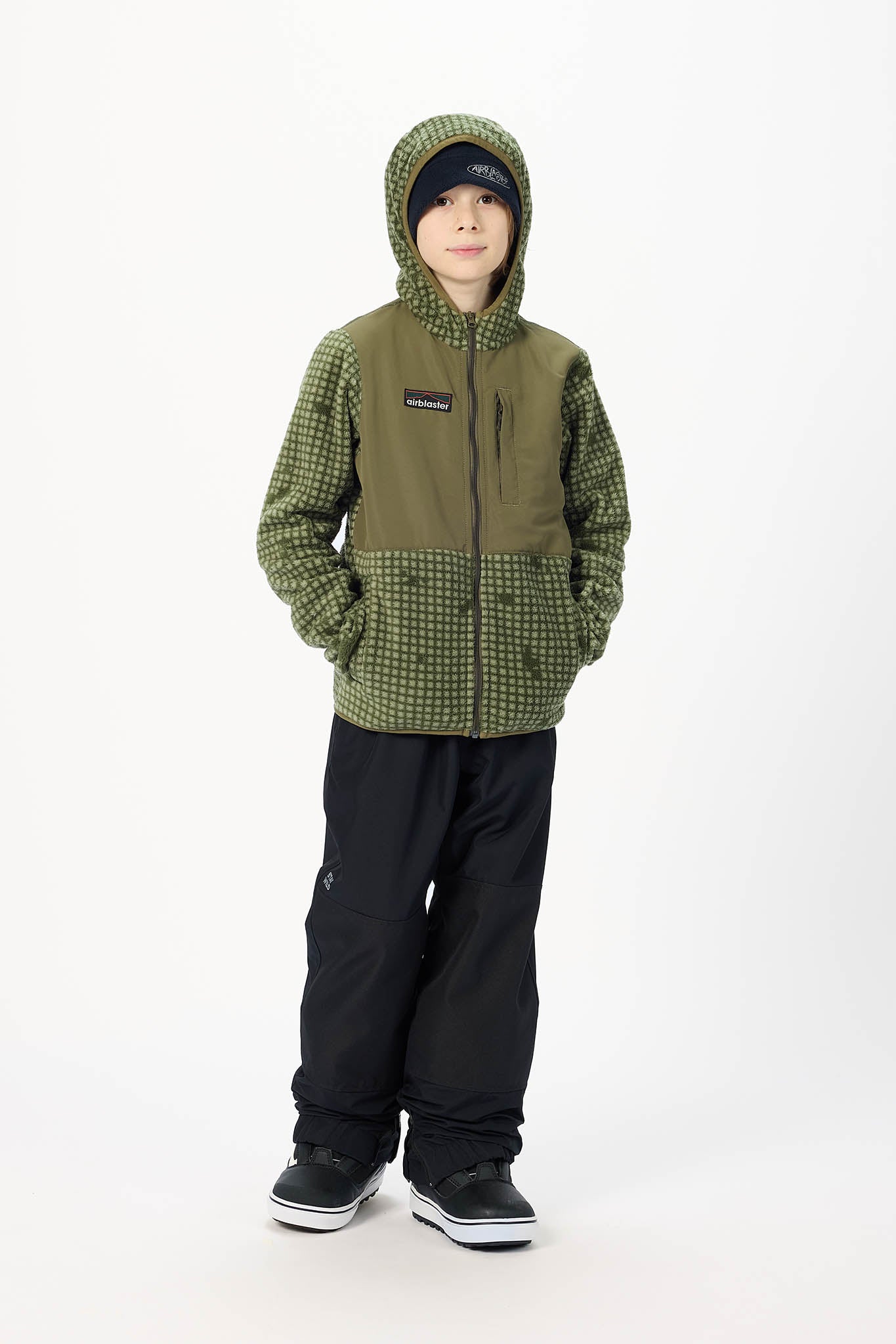 A child wearing a green and black patterned hooded jacket with black snow pants and black sneakers, standing against a plain white studio background.
