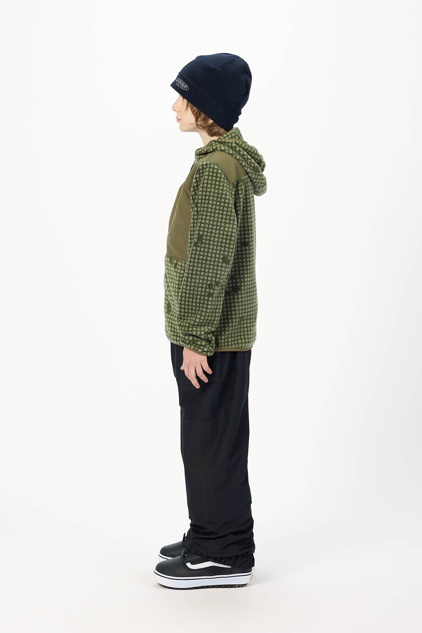 A child standing in profile wearing an olive green hooded jacket with a black-and-white check pattern on the sleeves and hood, black pants, a black beanie, and black sneakers with white soles, against a plain white studio background.