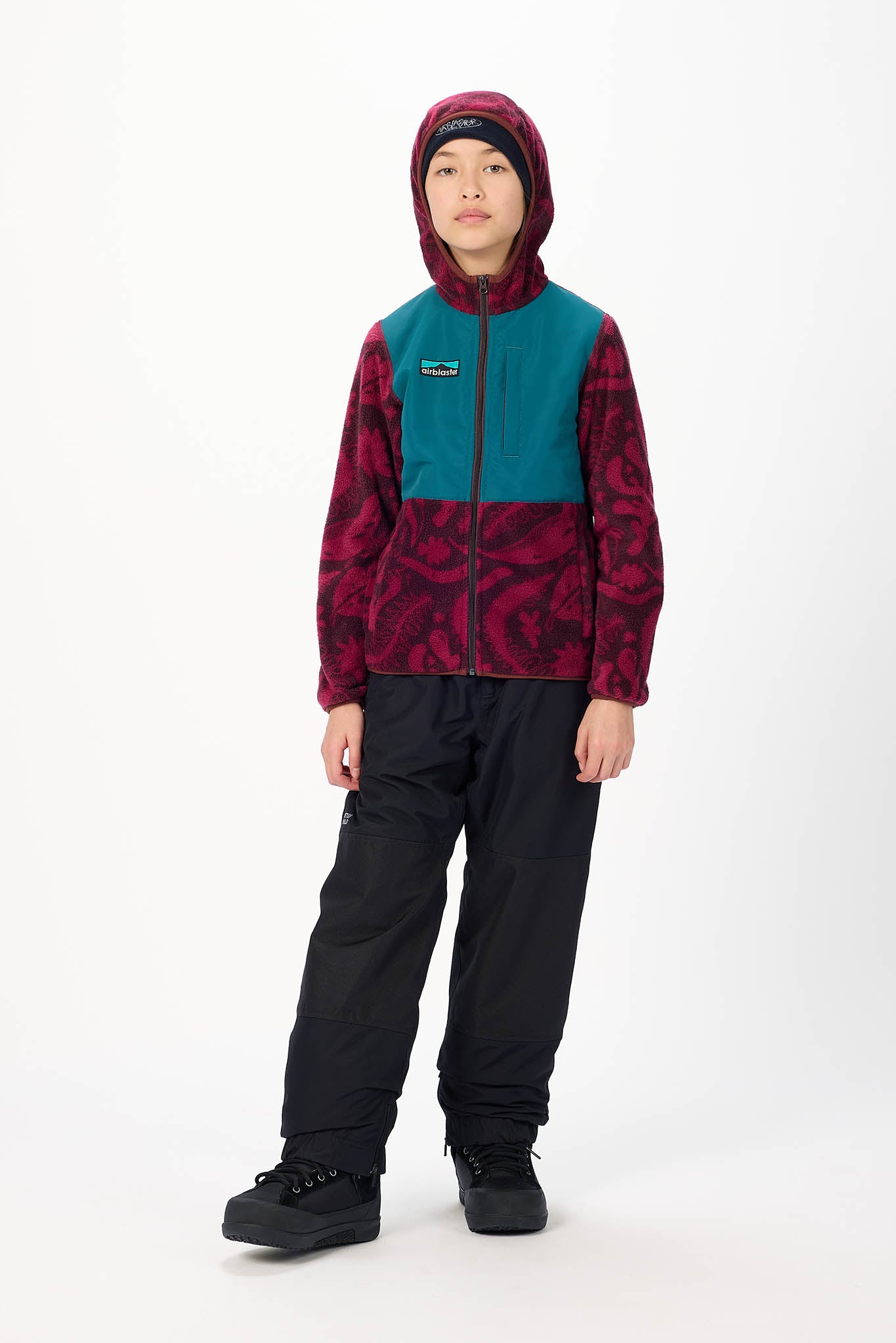 A child model wearing a Patagonia fleece jacket with a teal and burgundy patterned design, black snow pants, and black snow boots, standing against a white studio background.