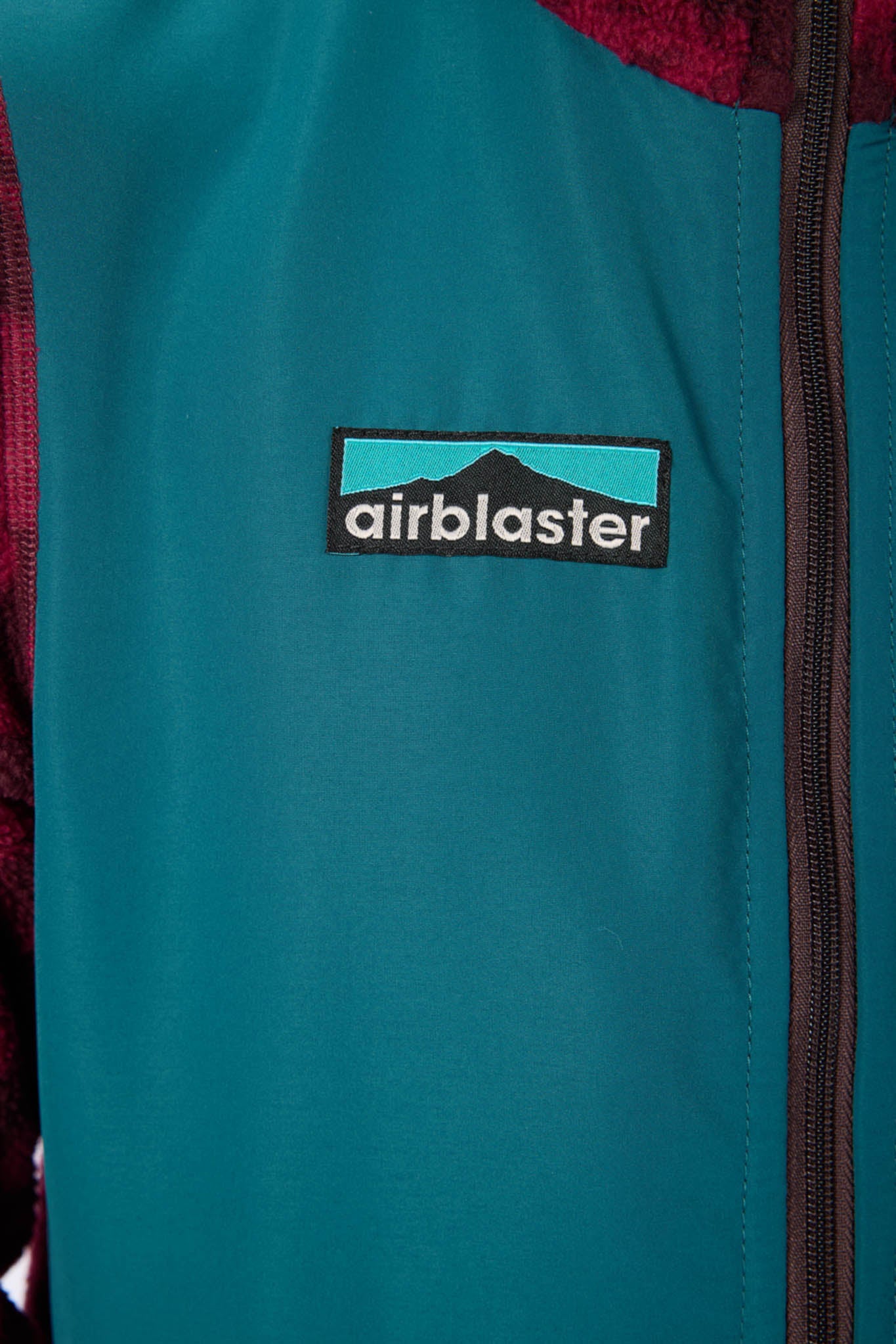 Close-up of a teal zip-front jacket with a visible Airblaster logo patch on the chest and dark zipper trim.