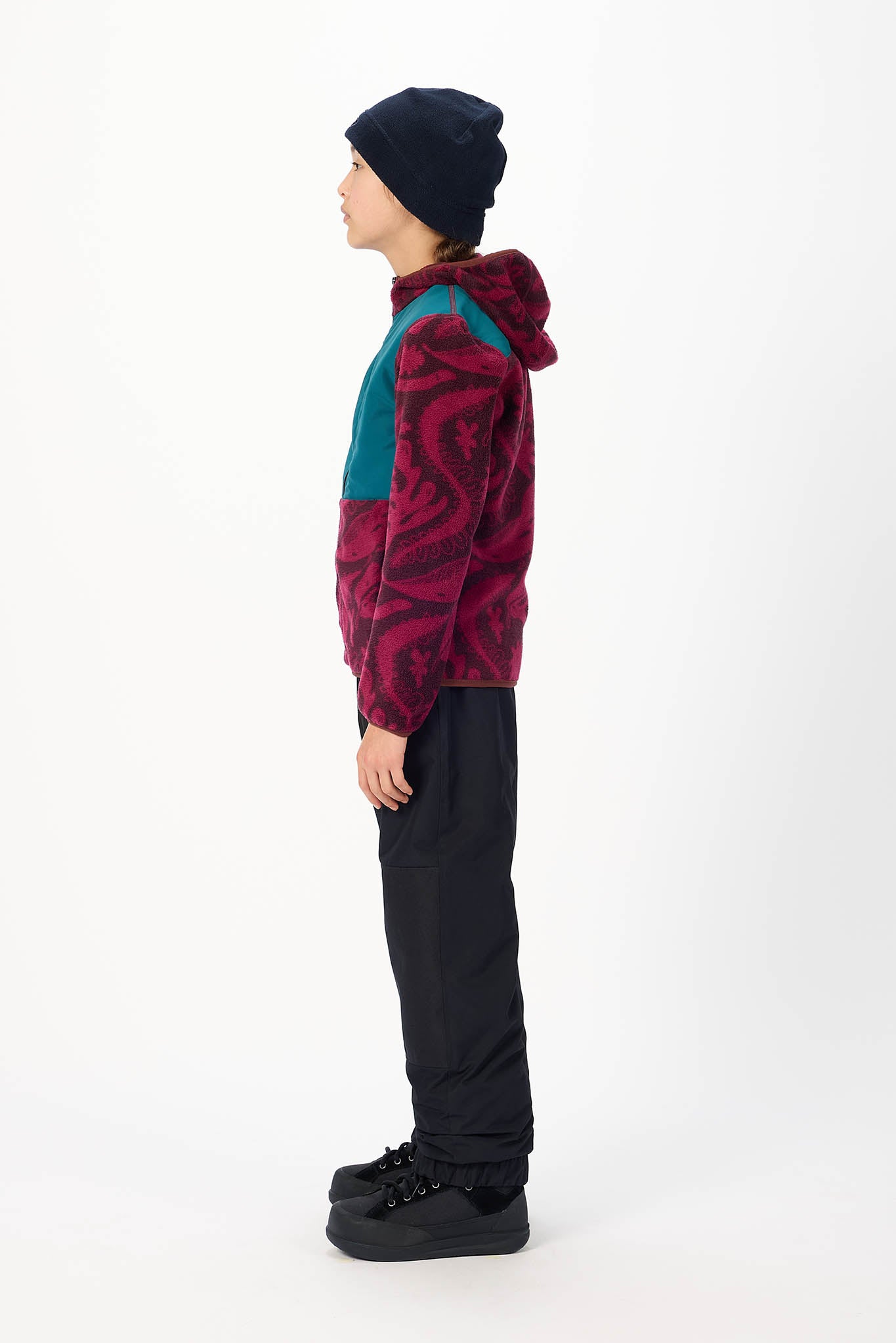 A child shown in profile wearing a dark navy beanie, a color-blocked fleece hoodie with a burgundy and teal pattern, black pants, and black platform sneakers against a plain white studio background.
