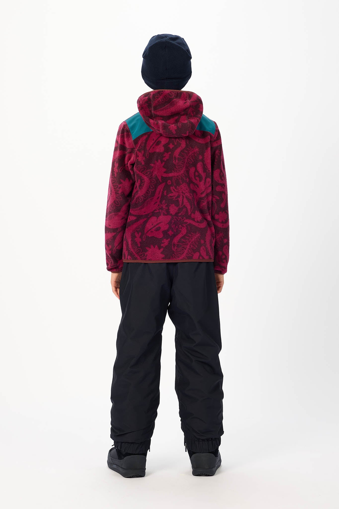 A child viewed from the back wearing a dark navy beanie, a patterned red and magenta hooded jacket with teal shoulder panels, and black snow pants against a plain white studio background.