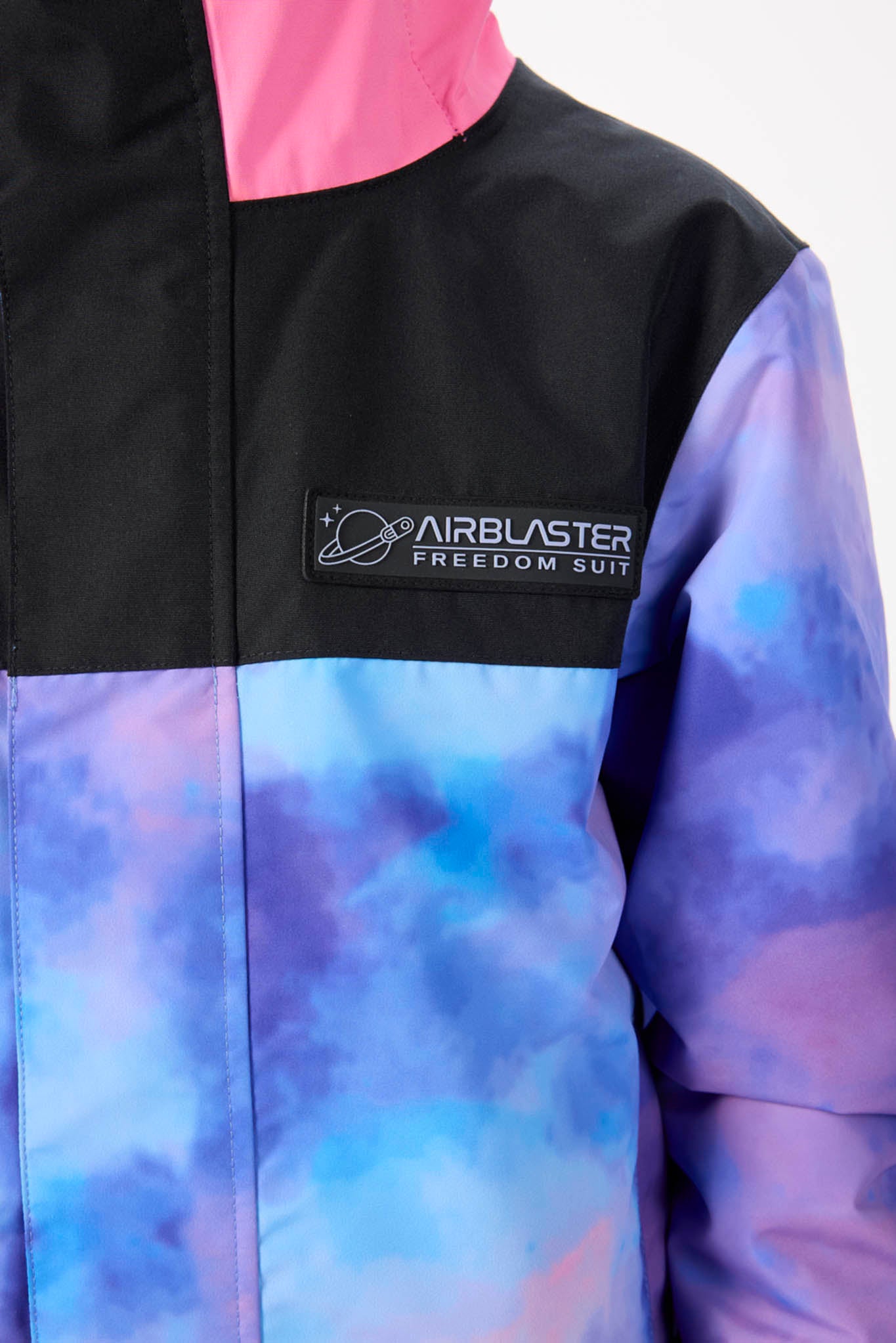 Close-up of a colorblock jacket featuring a black upper chest and collar with a pink lining, and a blue and purple tie-dye patterned lower body and sleeve. A black patch on the chest displays the 