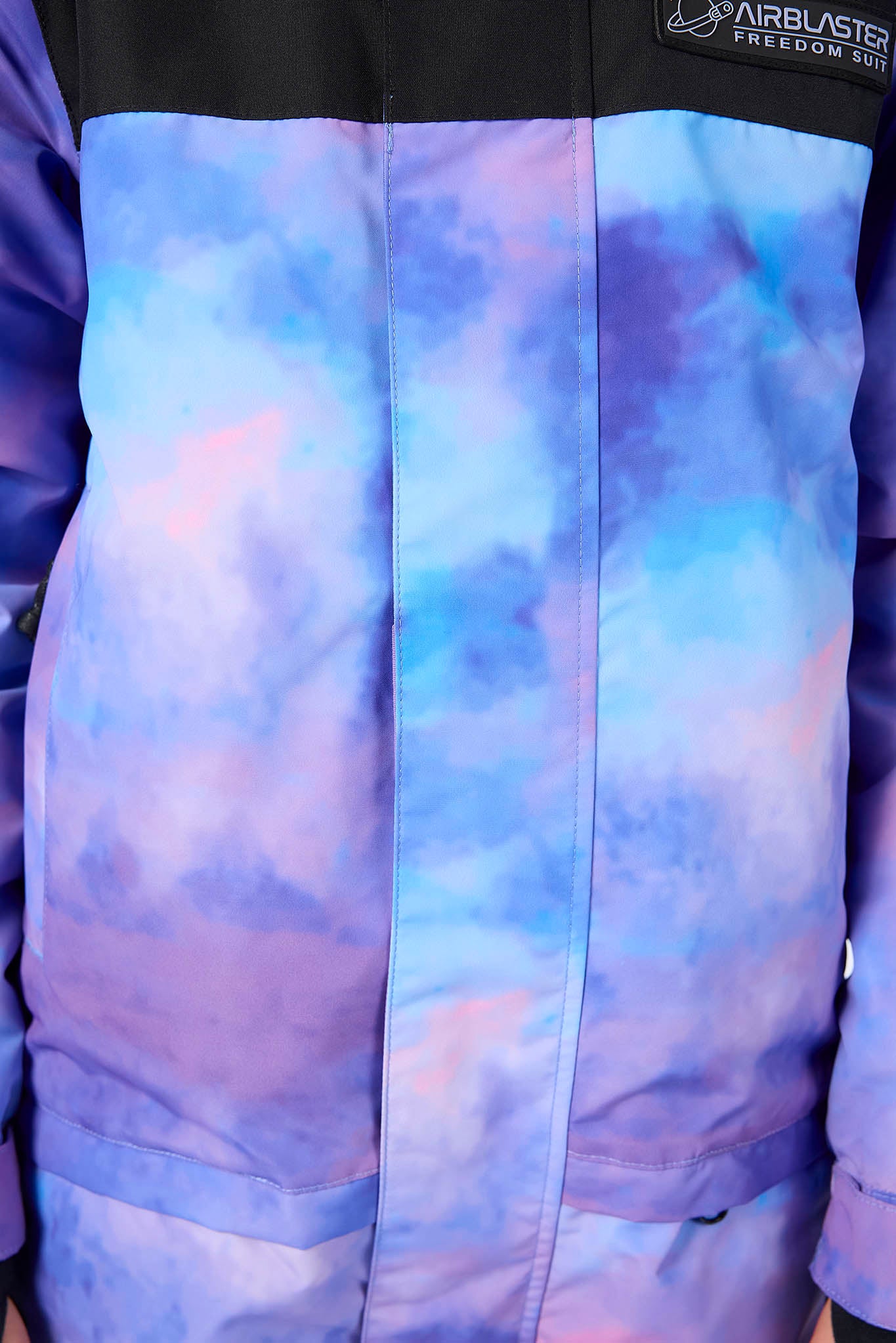 Close-up of a colorblock jacket featuring a black upper chest and collar with a pink lining, and a blue and purple tie-dye patterned lower body and sleeve. A black patch on the chest displays the 'Airblaster Freedom Suit' logo.