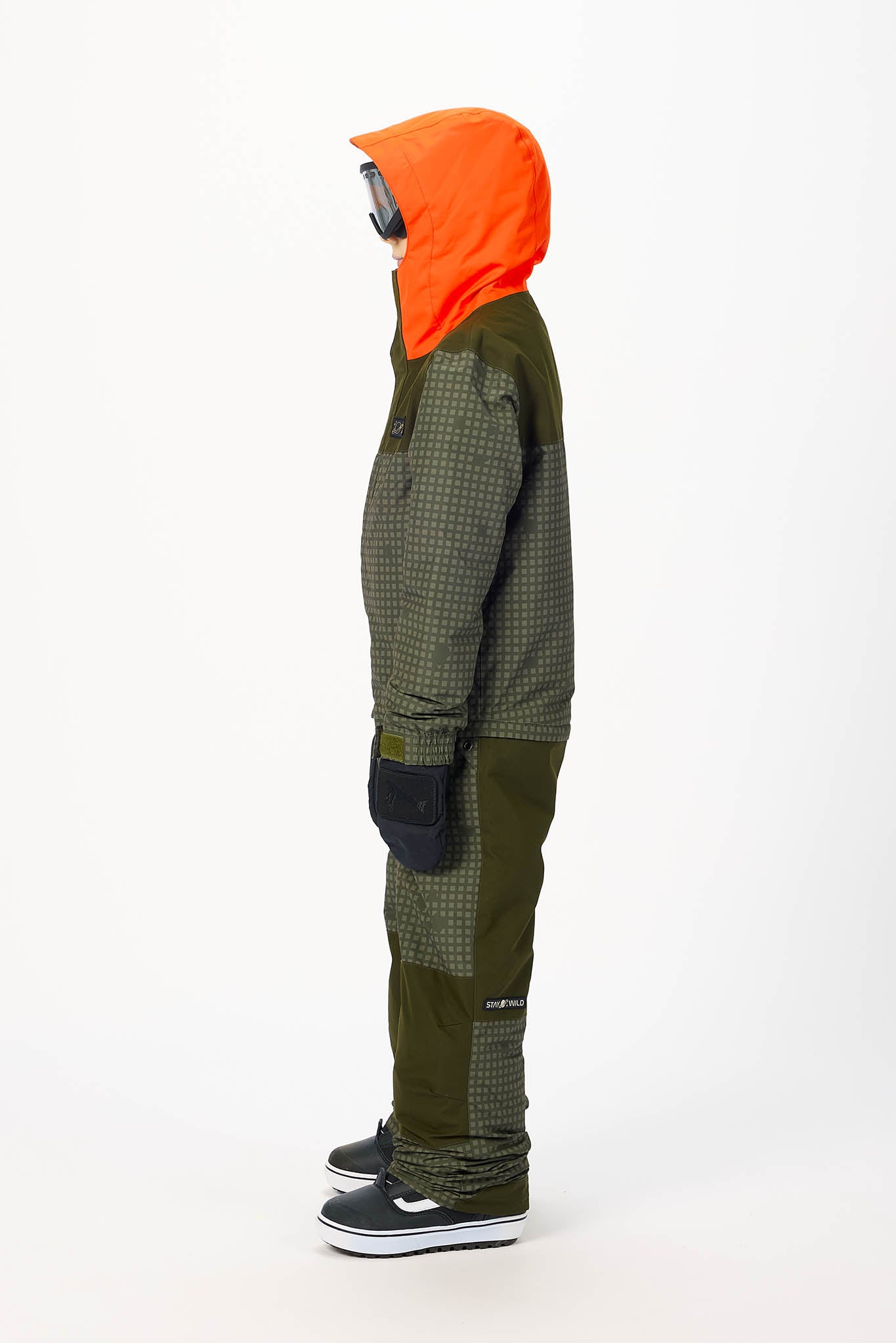 A side profile view of a person wearing a dark olive green and black checkered snowboard jacket with a bright orange hood, dark gloves, and black skate shoes against a plain white background.