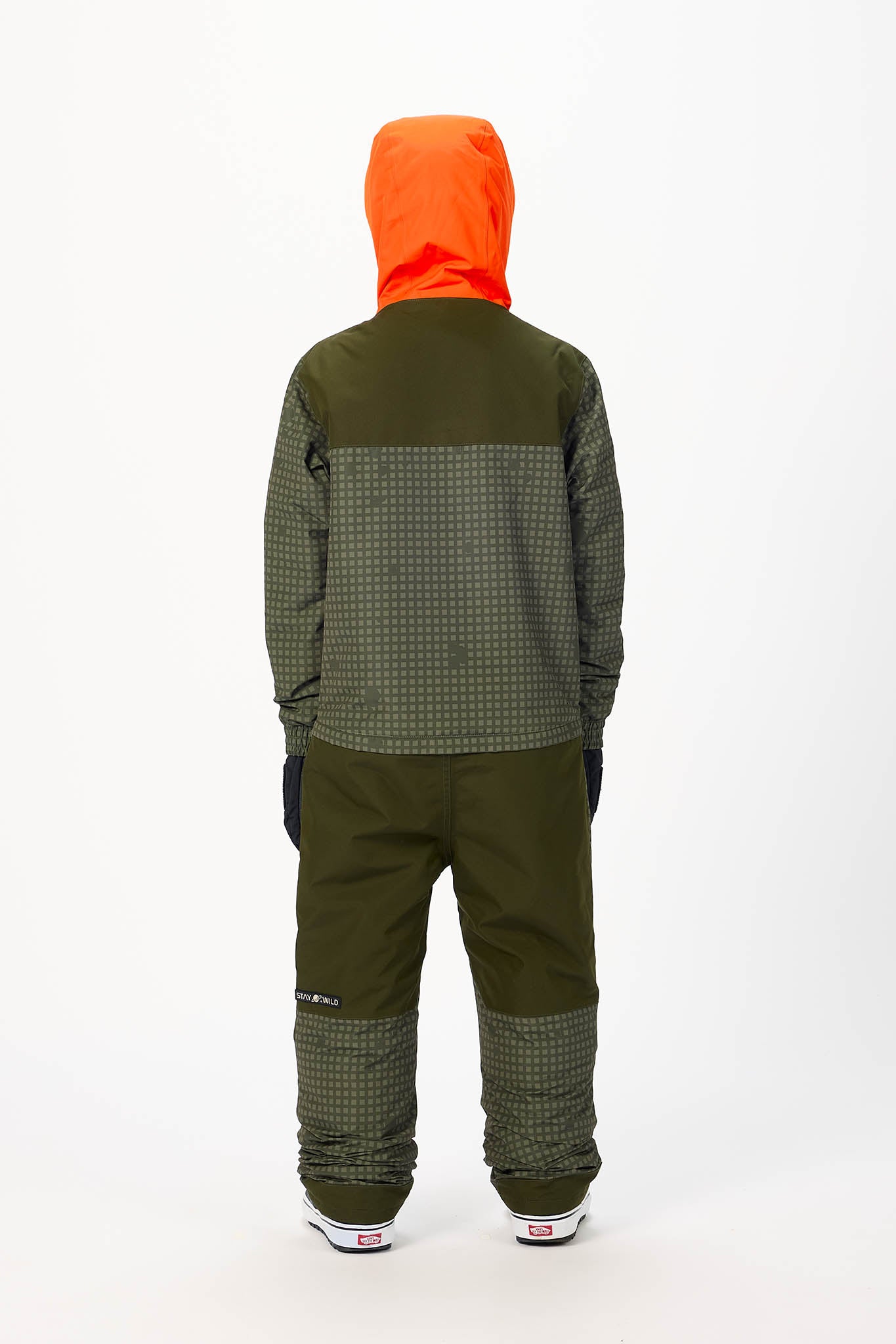 A child viewed from the back wearing a dark olive green and black patterned jacket with a bright orange hood, dark pants, and white sneakers on a white studio background.