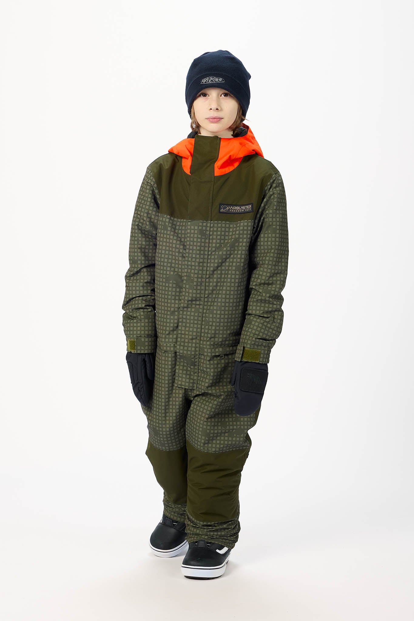 A child model wearing an olive green and black patterned one-piece snowsuit with a bright orange hood lining, a black beanie, and black sneakers, standing against a white studio background.