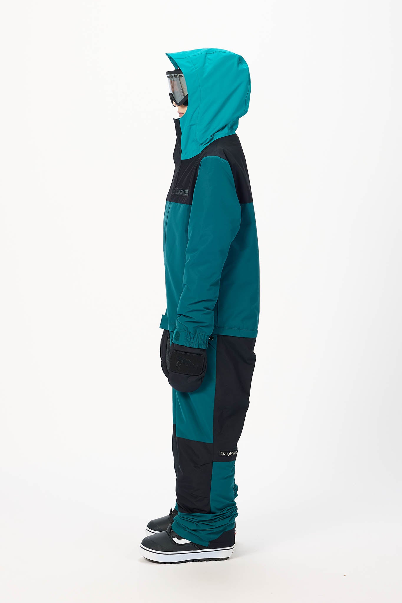 A person shown in profile wearing a teal and black hooded snowsuit with black snow boots, photographed against a plain white studio background.