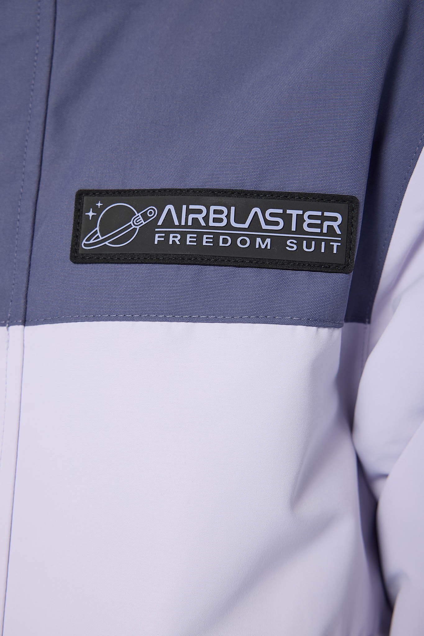 Close-up of a color-blocked jacket featuring a slate blue upper section and a white lower section, with a black rectangular patch on the chest reading 'AIRBLASTER FREEDOM SUIT'.
