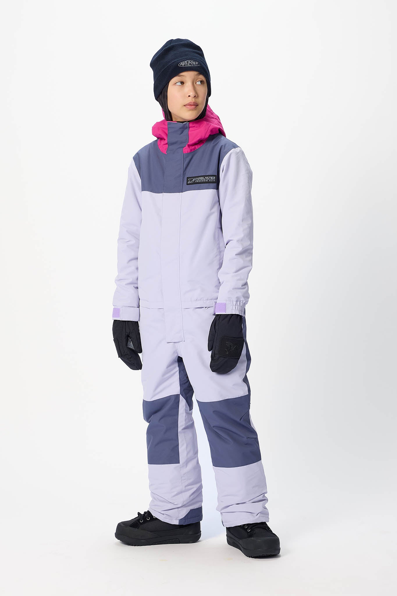 A child model wearing a Patagonia color-blocked snowsuit in light purple, slate blue, and pink, along with a navy beanie, black gloves, and black snow shoes, standing against a white studio background.