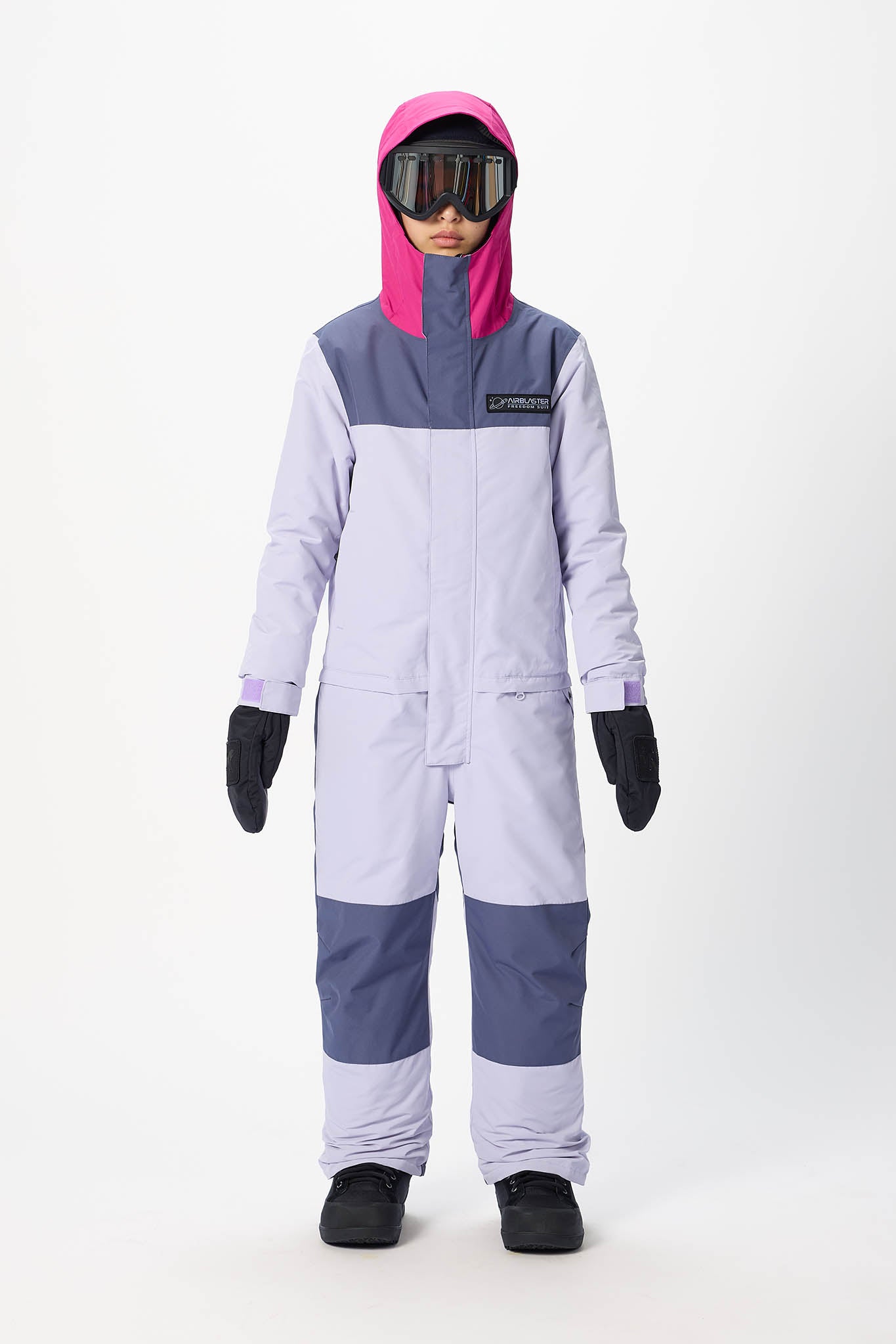 A child modeling a colorblock snowsuit with a light lavender body, slate blue panels, a bright pink hood, black gloves, and goggles against a plain white studio background.
