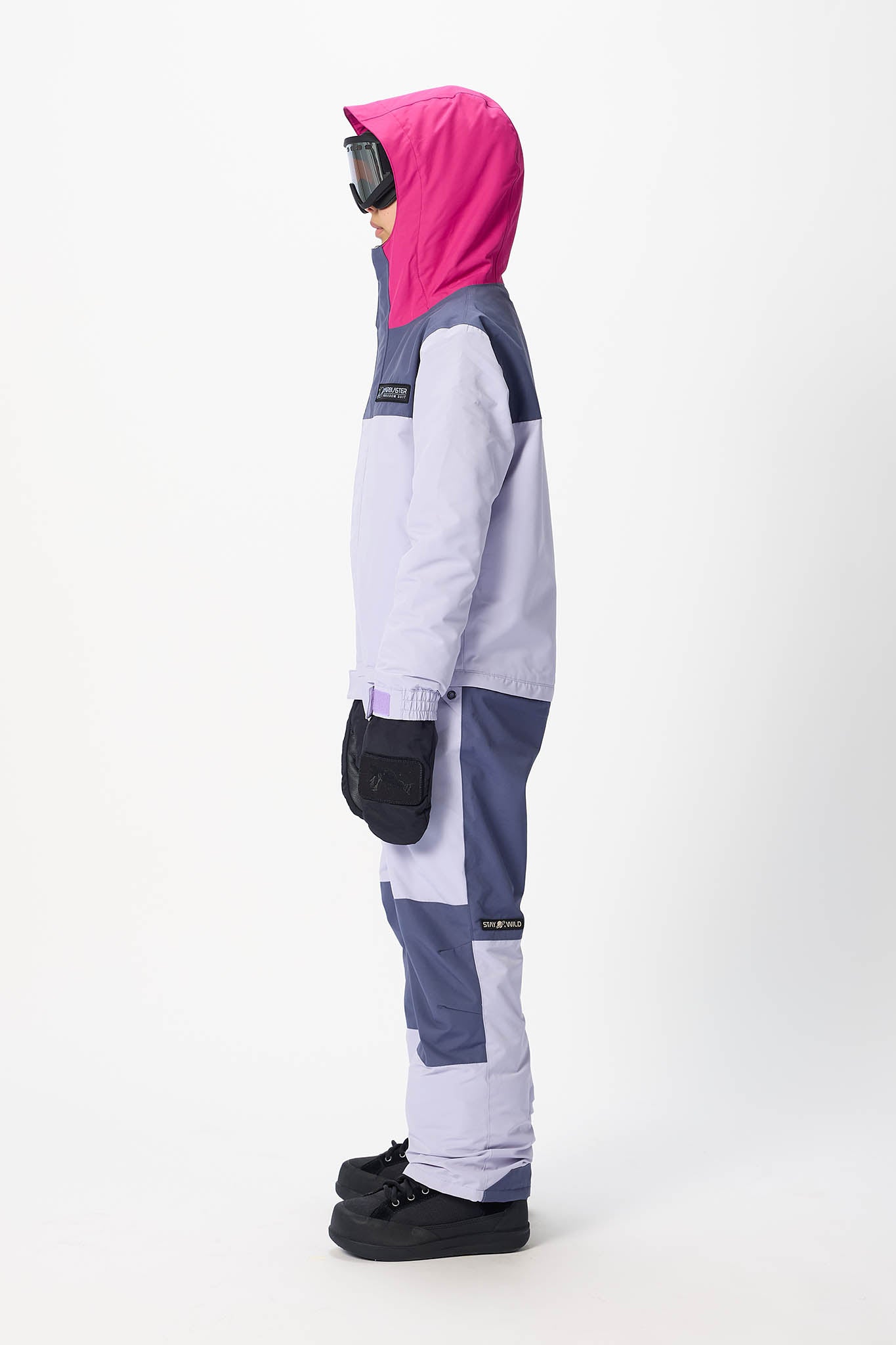 A child standing in profile wearing a color-blocked ski jacket with a bright pink hood, light gray and navy panels, black gloves, and black snow boots against a plain white studio background.