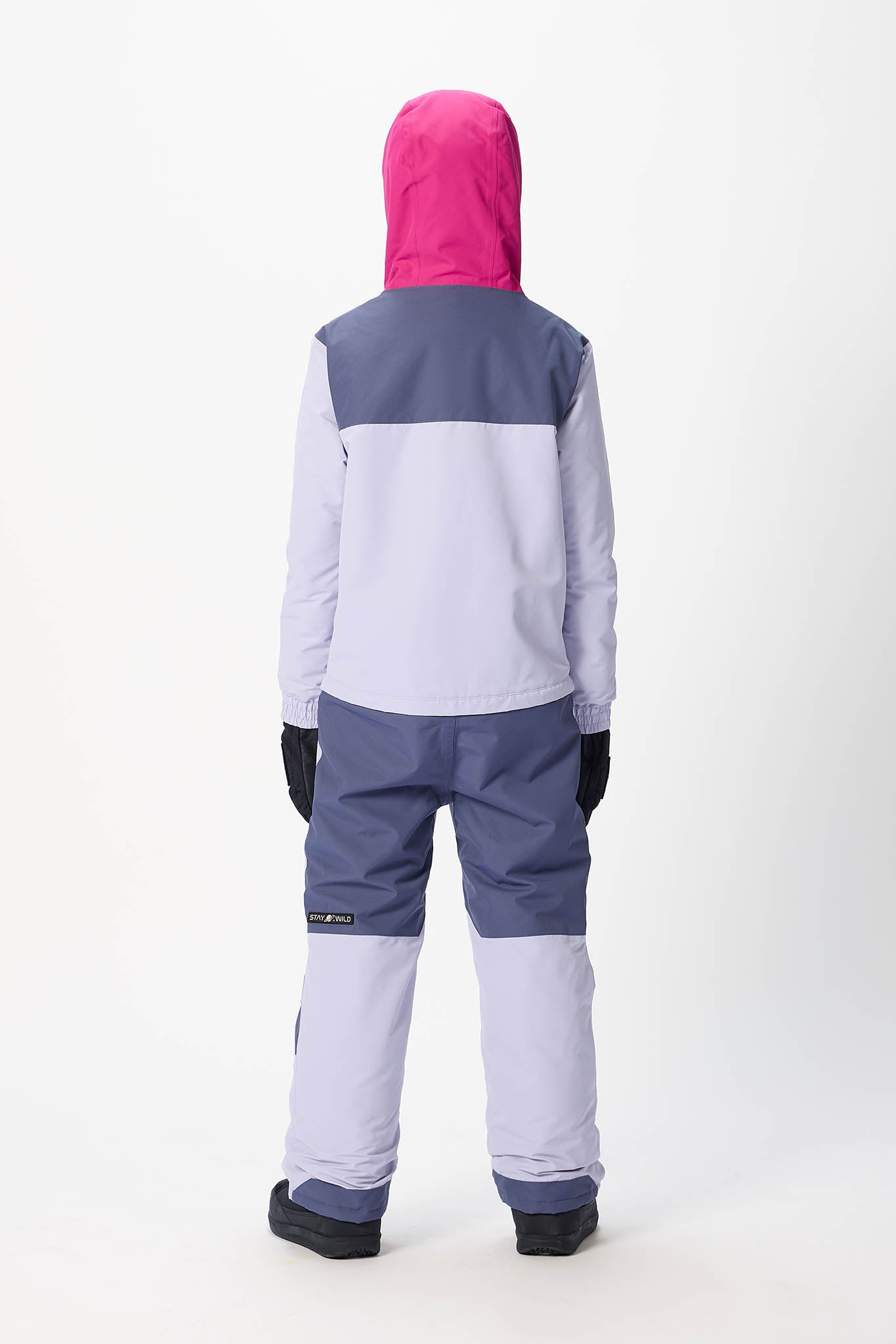 A child viewed from the back wearing a color-blocked snowsuit with a bright pink hood, a light lavender body, a dark slate blue yoke and lower legs, and black gloves, set against a plain white studio background.
