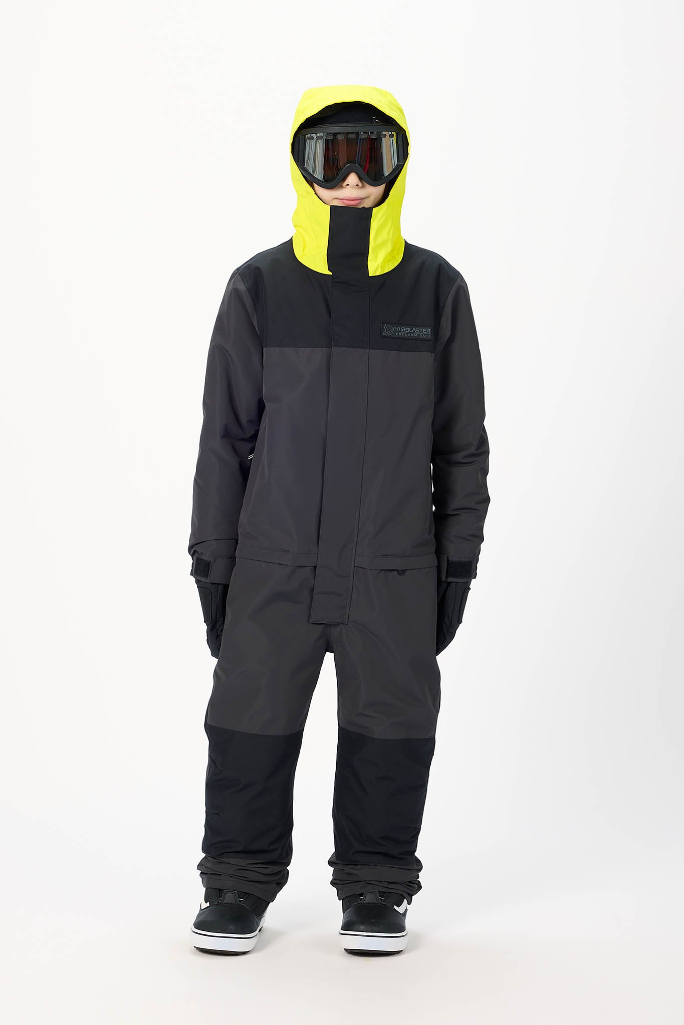 A child modeling a dark gray and black snowsuit with a bright yellow hood, black gloves, and ski goggles against a plain white studio background.