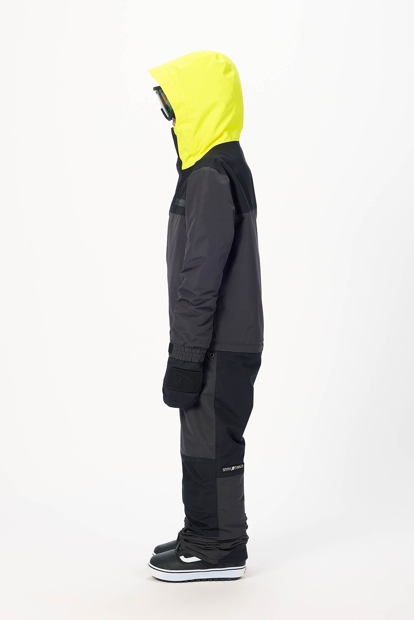 A child wearing a dark gray and black ski jacket with a bright neon yellow hood, black ski pants, and black sneakers, shown in profile against a plain white studio background.