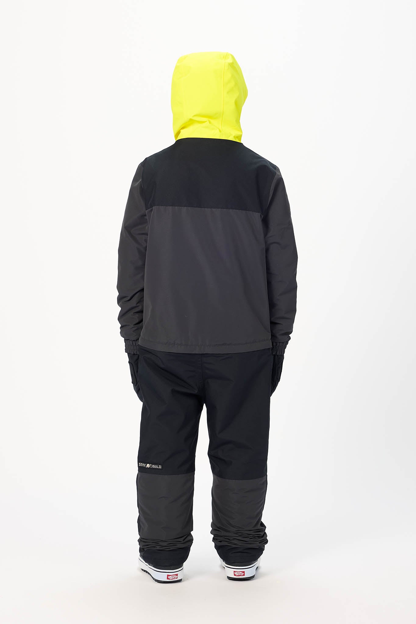 A child viewed from the back wearing a color-blocked hooded jacket with a bright yellow hood, dark gray upper back, and black lower body, paired with dark pants and white sneakers, against a plain white studio background.