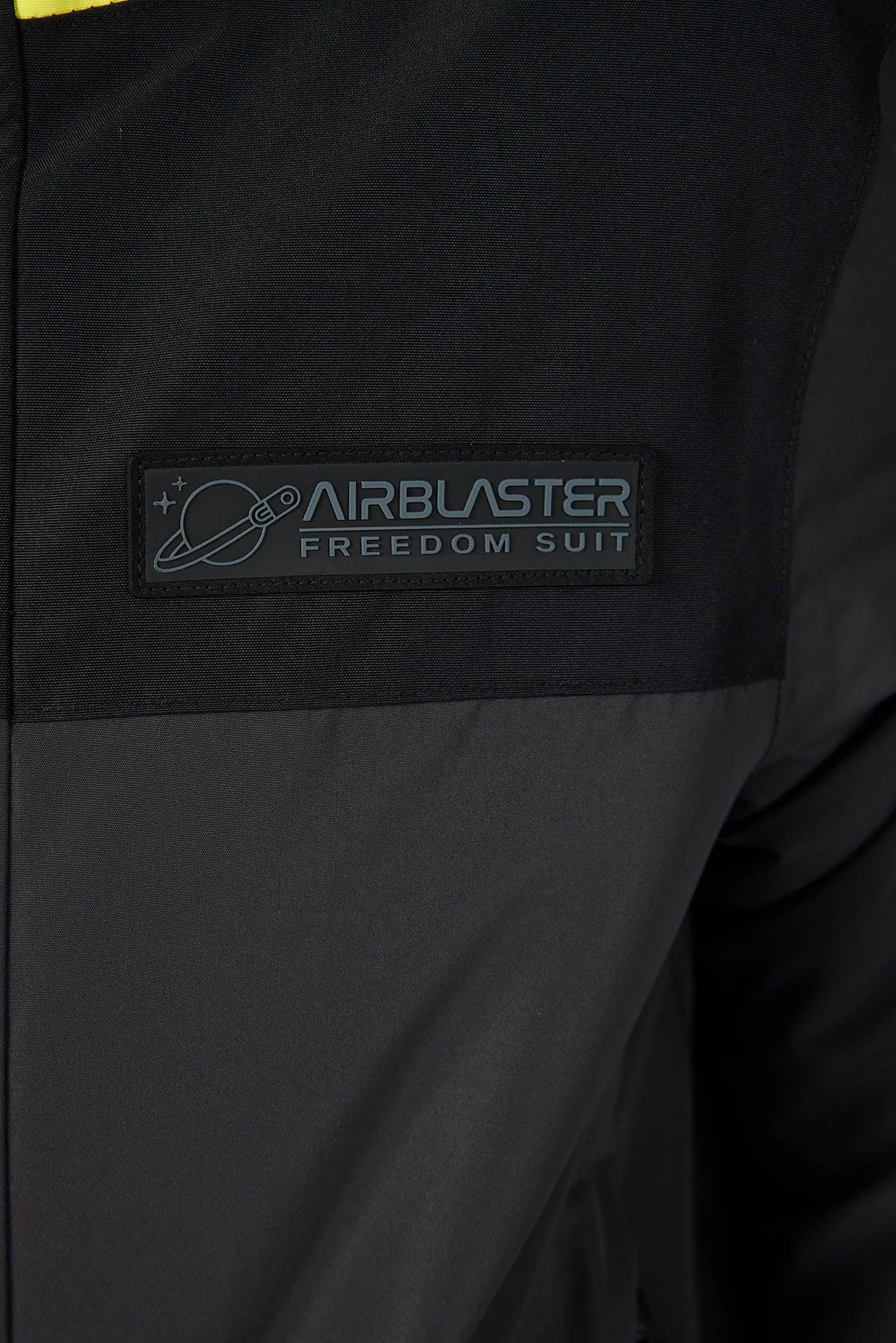 Close-up of a black jacket or suit with a rectangular patch reading 'AIRBLASTER FREEDOM SUIT' and a small planet graphic, shown against a dark fabric background.