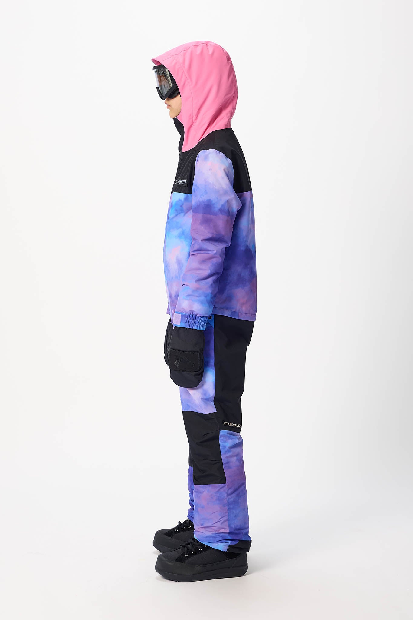 A child model shown in profile wearing a hooded ski jacket and matching pants with a purple and blue galaxy tie-dye pattern and black panels, against a plain white studio background.