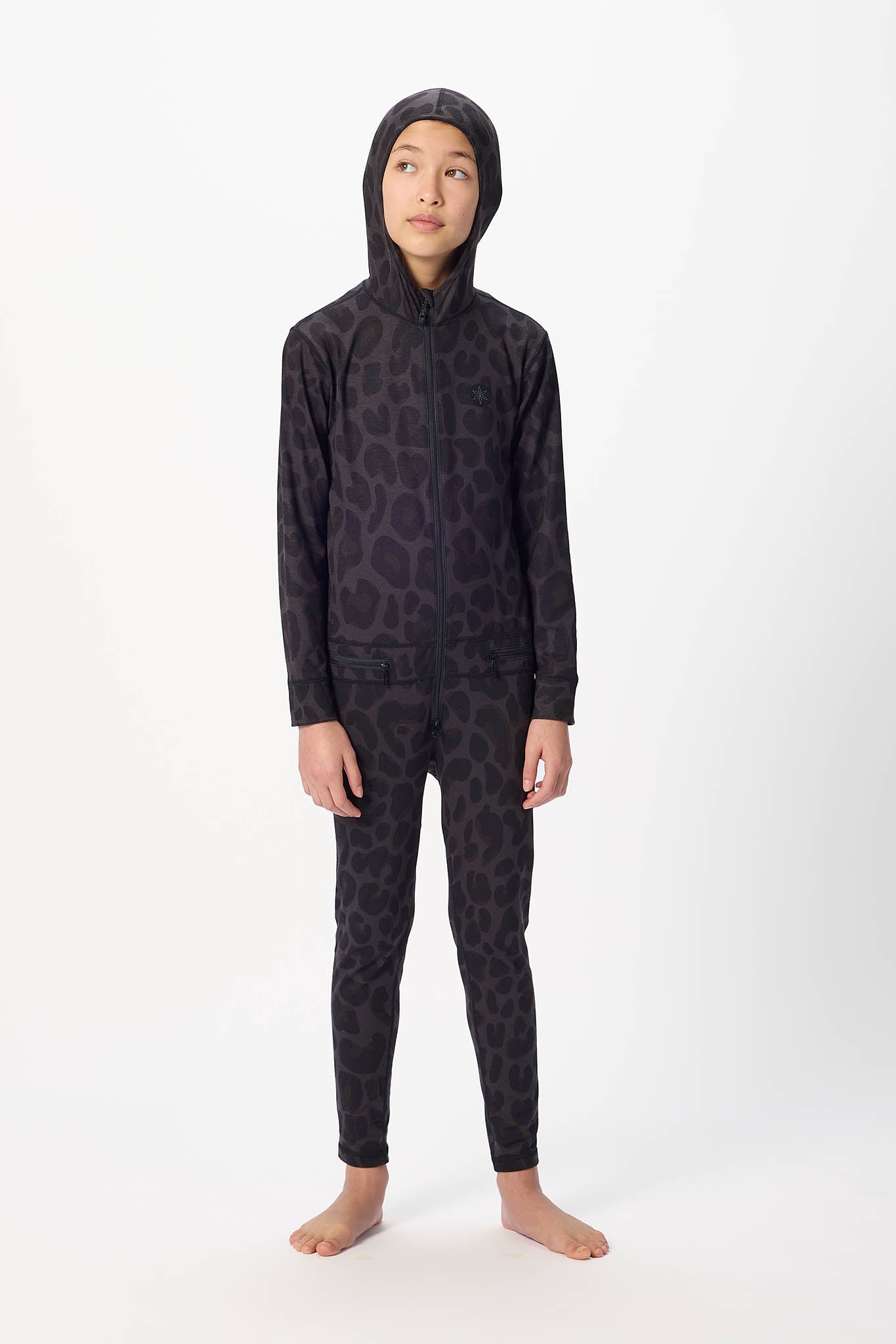 A child modeling a black full-body hooded one-piece jumpsuit with a subtle tonal leopard-like pattern, standing against a plain white studio background.