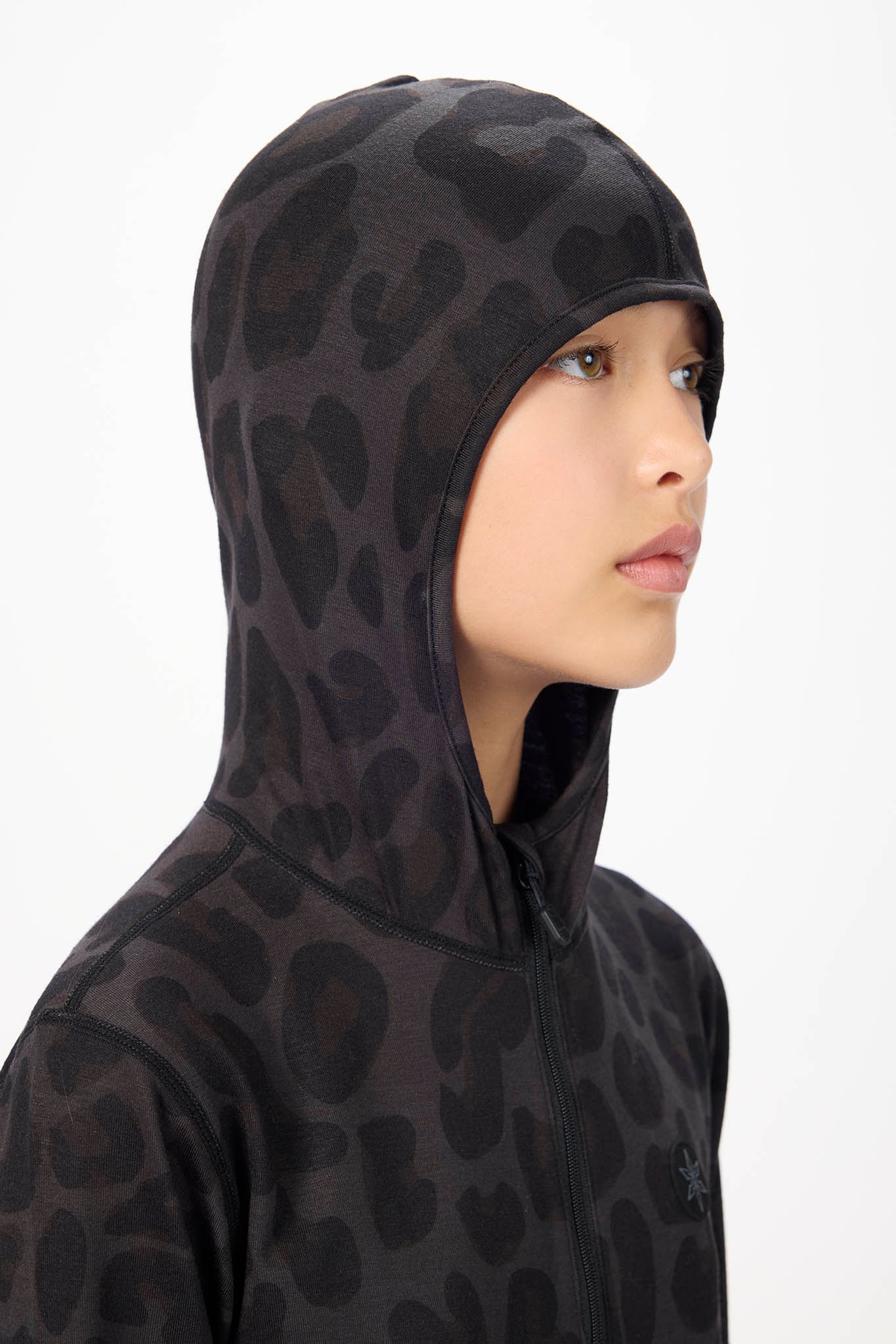 A close-up side profile of a person wearing a black hooded sweatshirt with a dark gray tonal leopard print pattern, set against a plain white background.