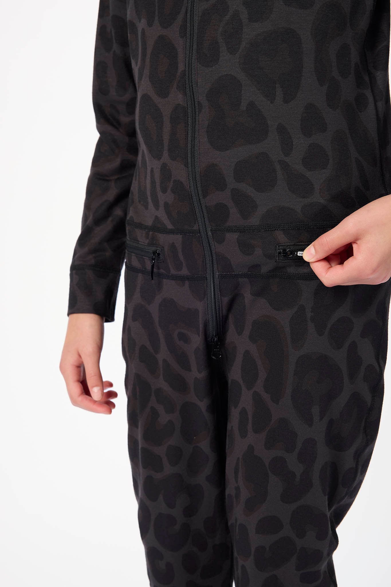 A close-up of a person wearing a black long-sleeve zip-front jumpsuit or one-piece with a tonal leopard-like spot pattern. A hand is shown pulling a zipper near the waist.