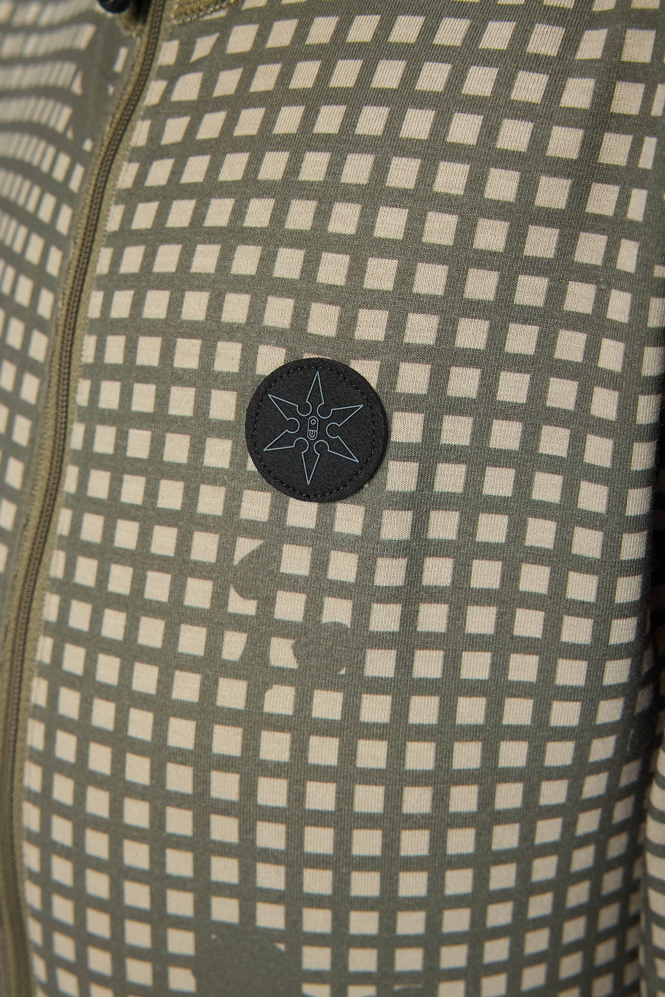 Close-up of a patterned jacket or fabric panel with a repeating small square grid in muted olive green and off-white, plus a dark circular patch with a light geometric star-like emblem near the center.
