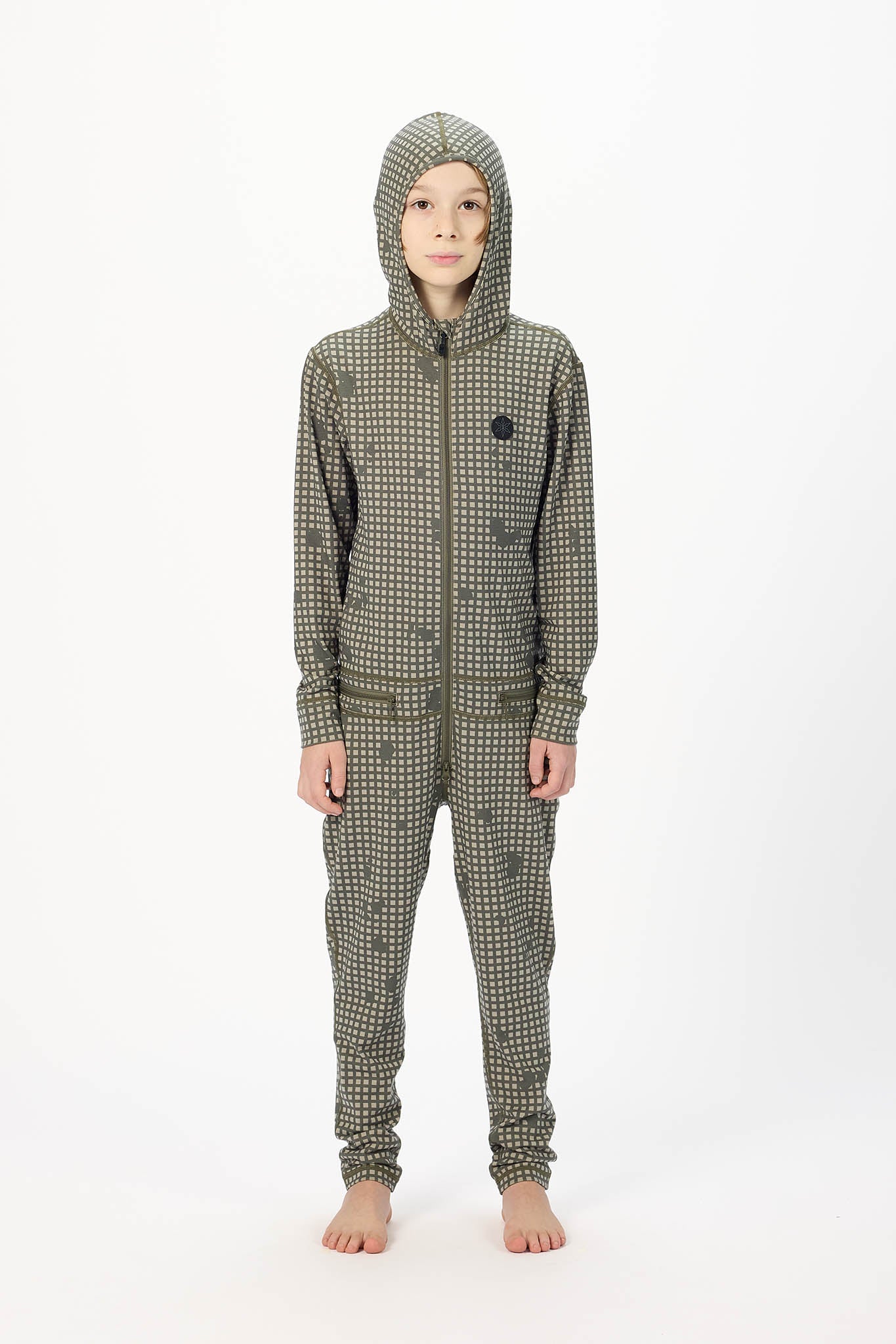 A child standing barefoot against a plain white studio background, wearing a hooded one-piece jumpsuit with a small-scale dark pattern over an olive green base.