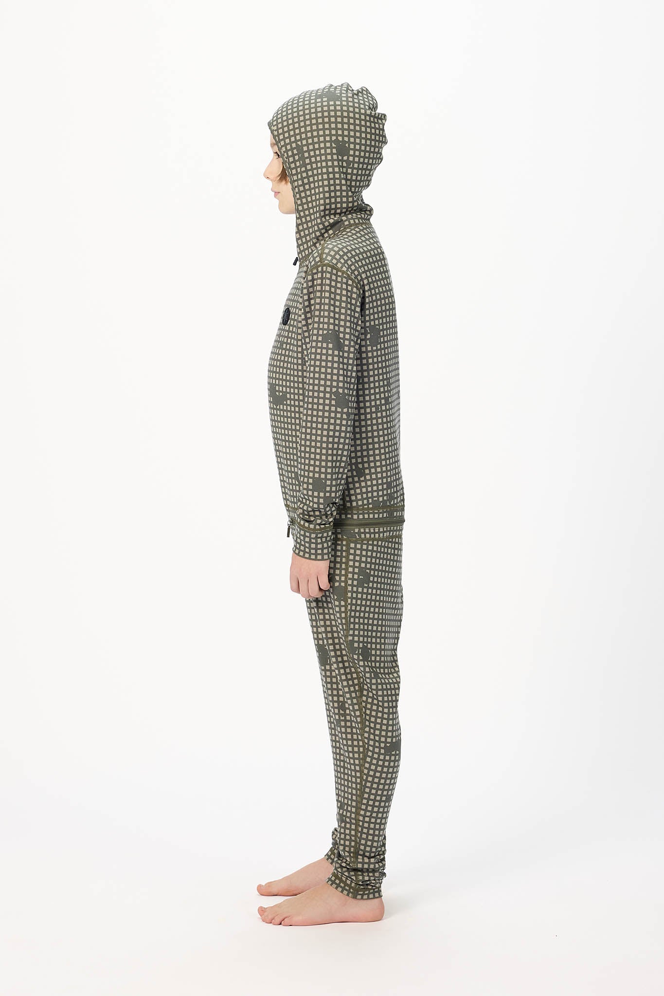A child standing in profile wearing a hooded one-piece outfit with an allover small-scale check pattern in olive green and white, photographed against a plain white studio background.