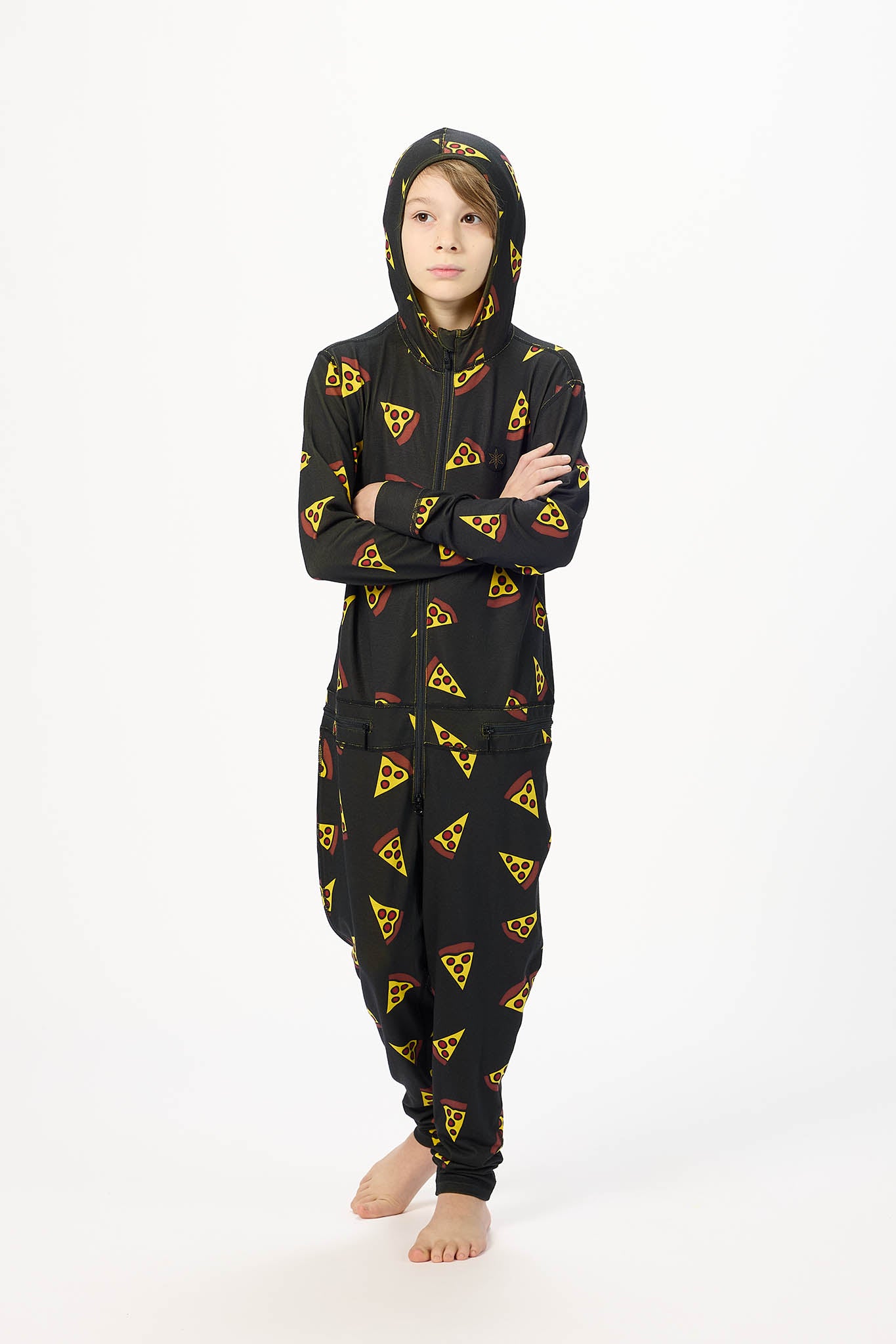 A child wearing a black hooded one-piece pajama or kigurumi with an all-over pizza slice print, standing against a plain white studio background with arms crossed.