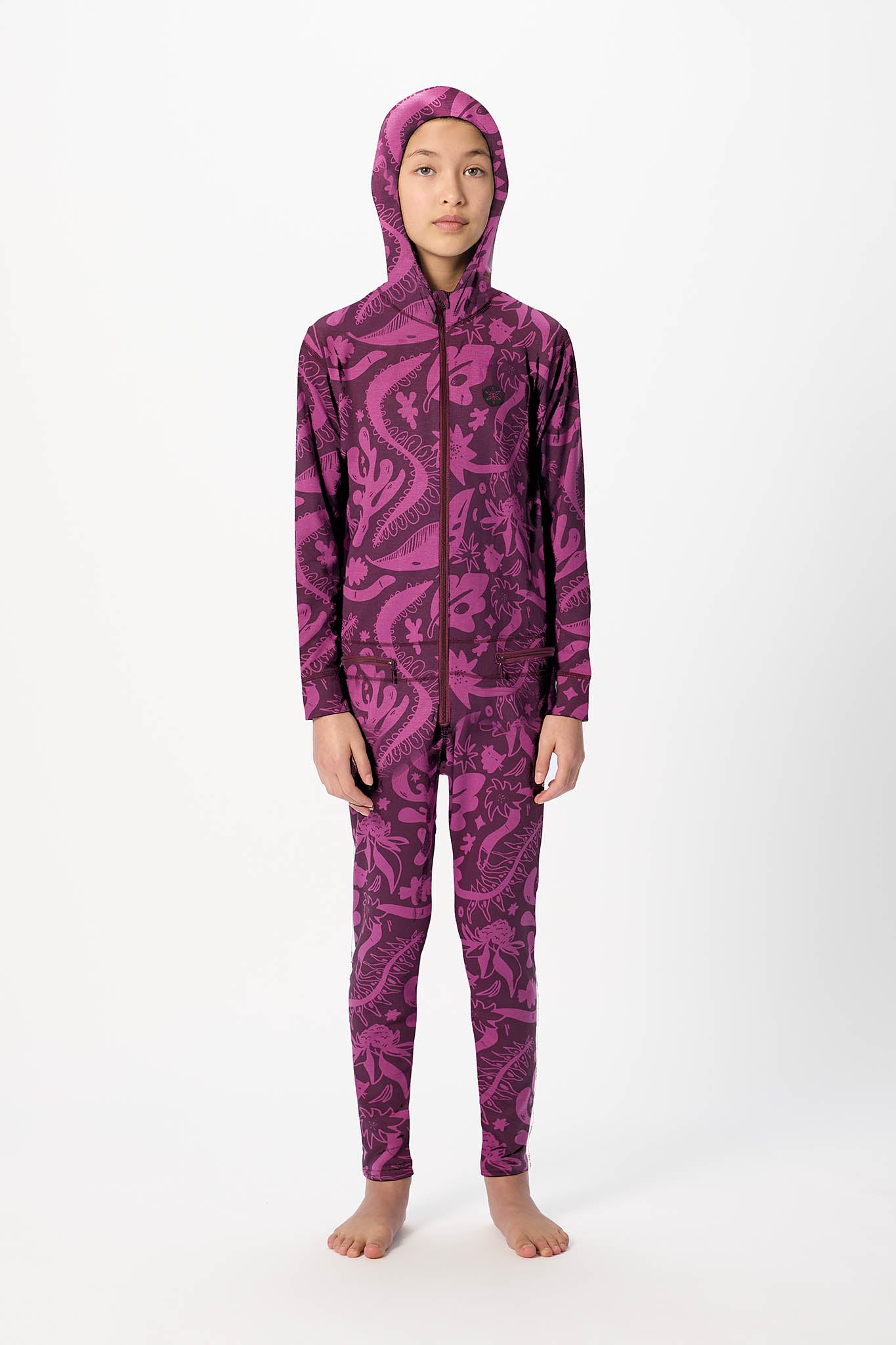 A child model wearing a full-body hooded one-piece swimsuit or wetsuit with an all-over dark paisley pattern on a bright magenta-pink base, standing against a plain white studio background.