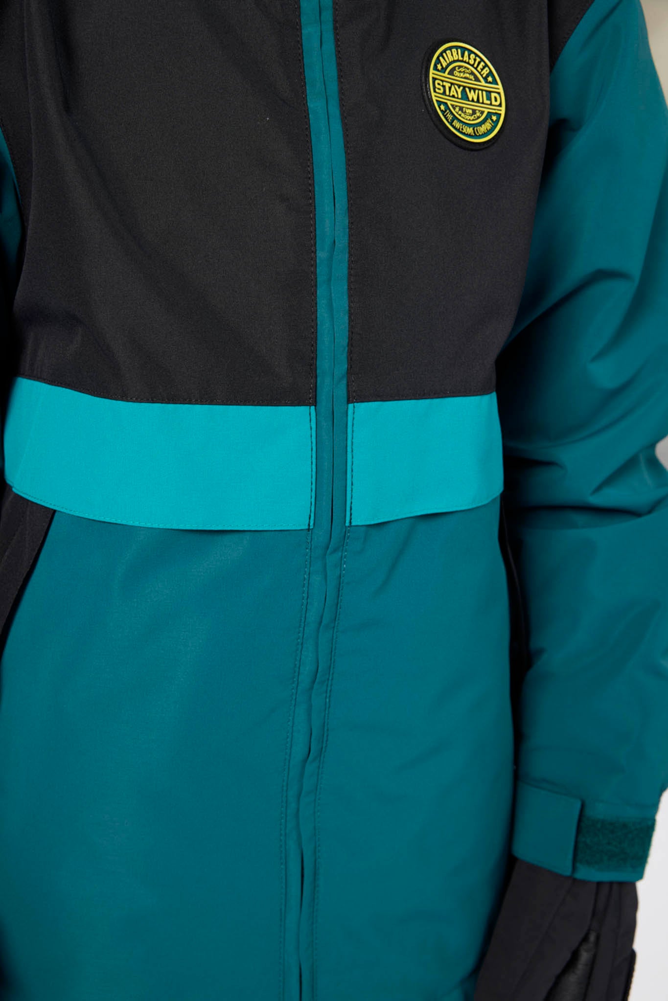 Close-up of a colorblock ski jacket featuring a black upper section, a teal midsection with a horizontal stripe, and a yellow circular 'Arctic Exploration' logo on the left chest.