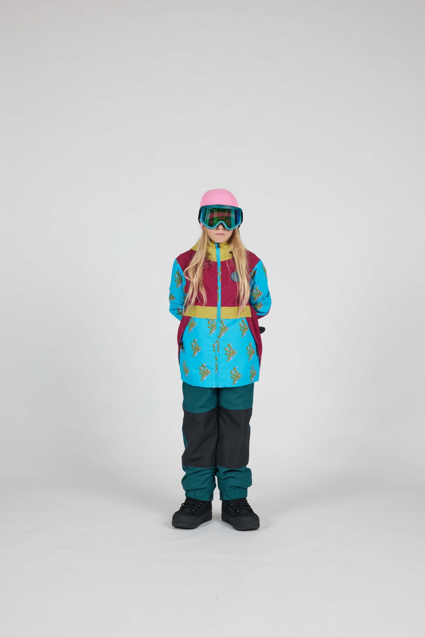 A child model wearing a colorful ski outfit consisting of a turquoise and magenta patterned jacket, dark teal pants, a pink helmet, and goggles, standing against a neutral gray studio background.