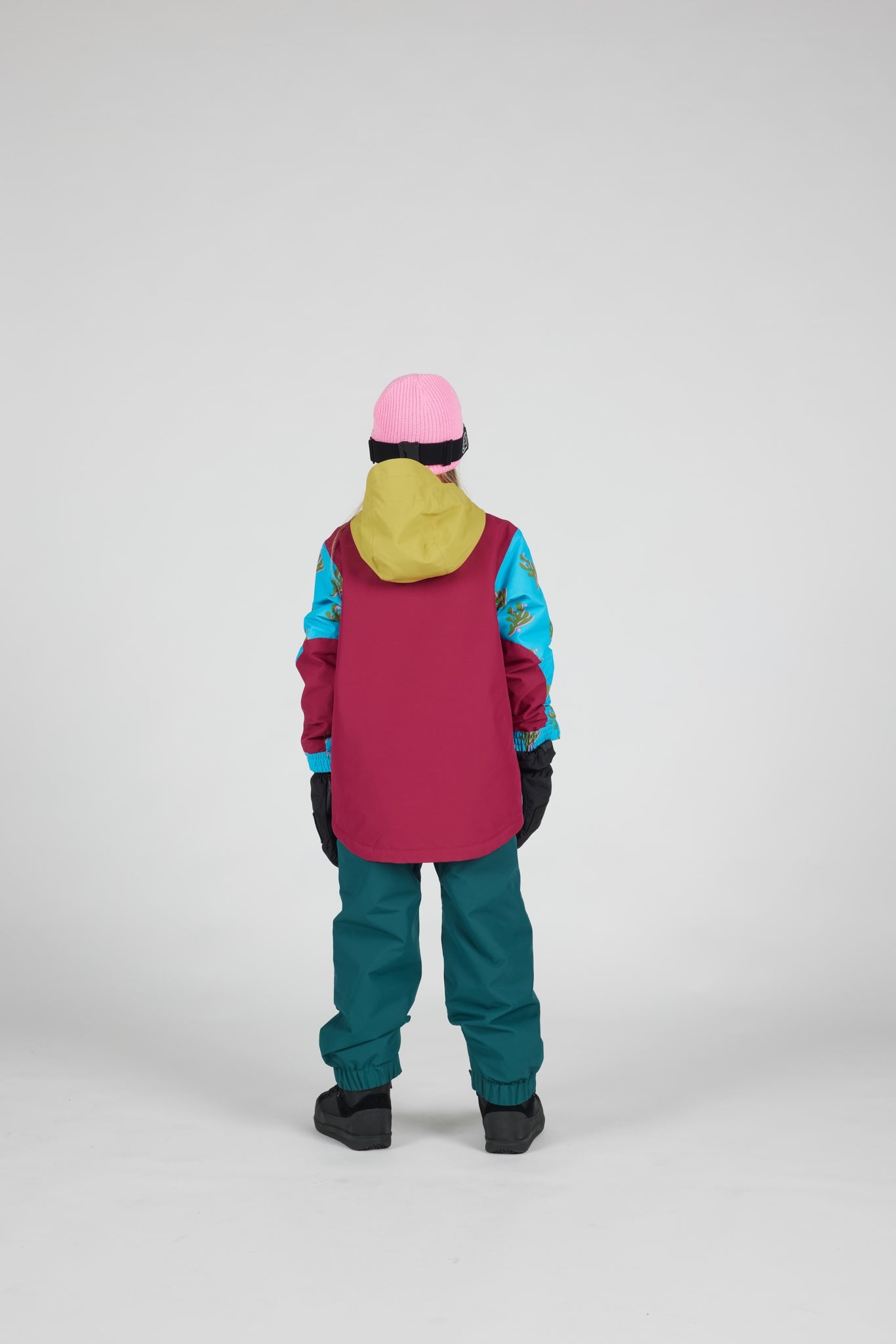 A child standing with their back to the camera, wearing a multi-colored ski jacket with a magenta body, yellow hood, blue sleeves, teal pants, a pink beanie, and black gloves against a light gray studio background.