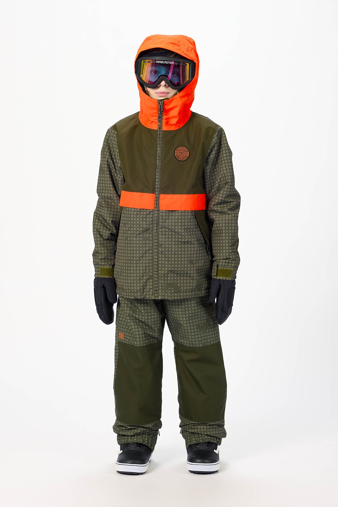 A child wearing a dark olive green and orange snowboard suit with a patterned texture, black gloves, and goggles, standing against a plain white studio background.