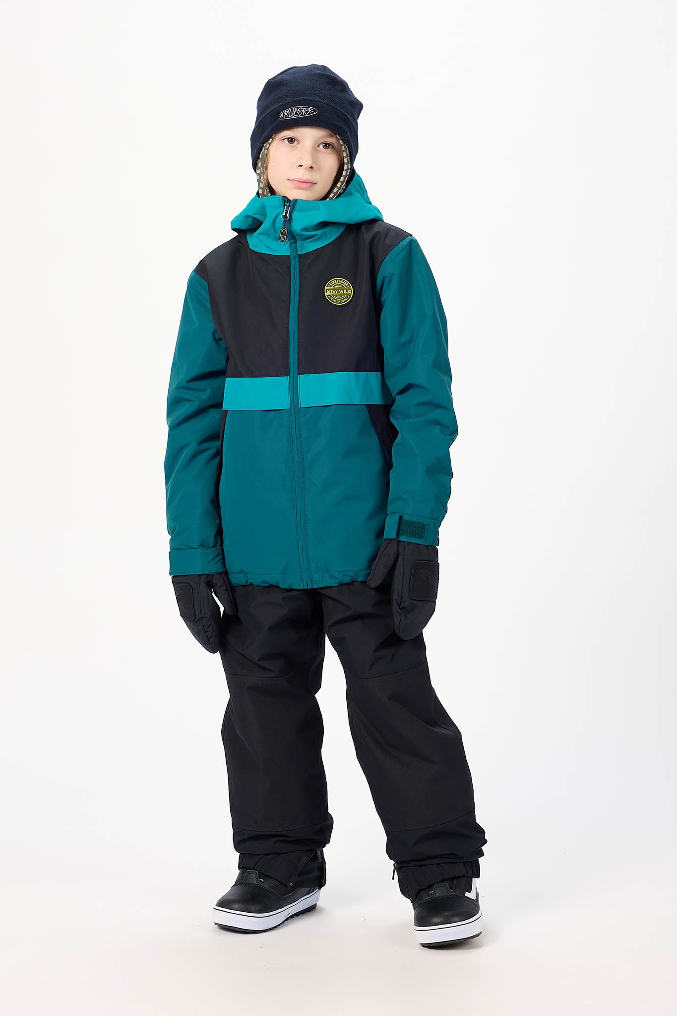 A child wearing a color-blocked ski jacket in teal and black, black snow pants, a dark navy beanie, and black snow boots, standing against a plain white studio background.