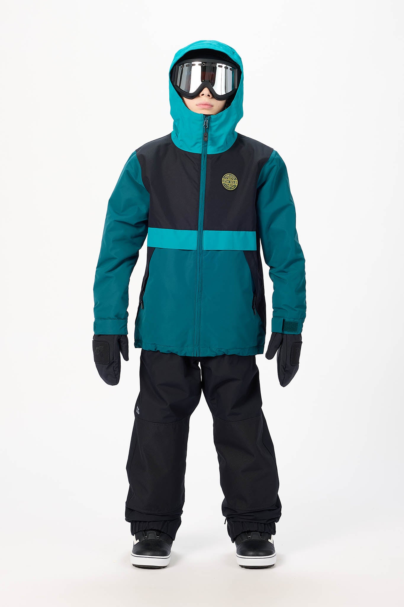 A child wearing a colorblock snowboard jacket in teal and black, black snow pants, black gloves, and goggles, standing against a plain white studio background.