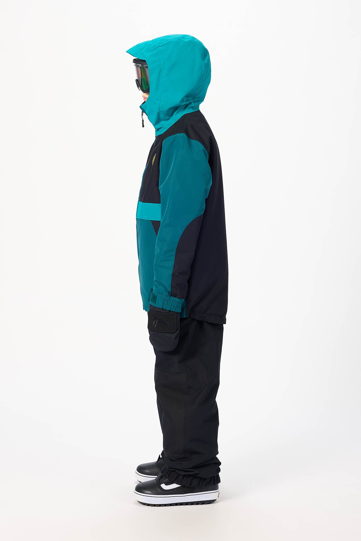 A child model shown in profile wearing a color-blocked ski jacket with a bright teal hood and teal chest panel, paired with black pants and black sneakers, against a plain white studio background.