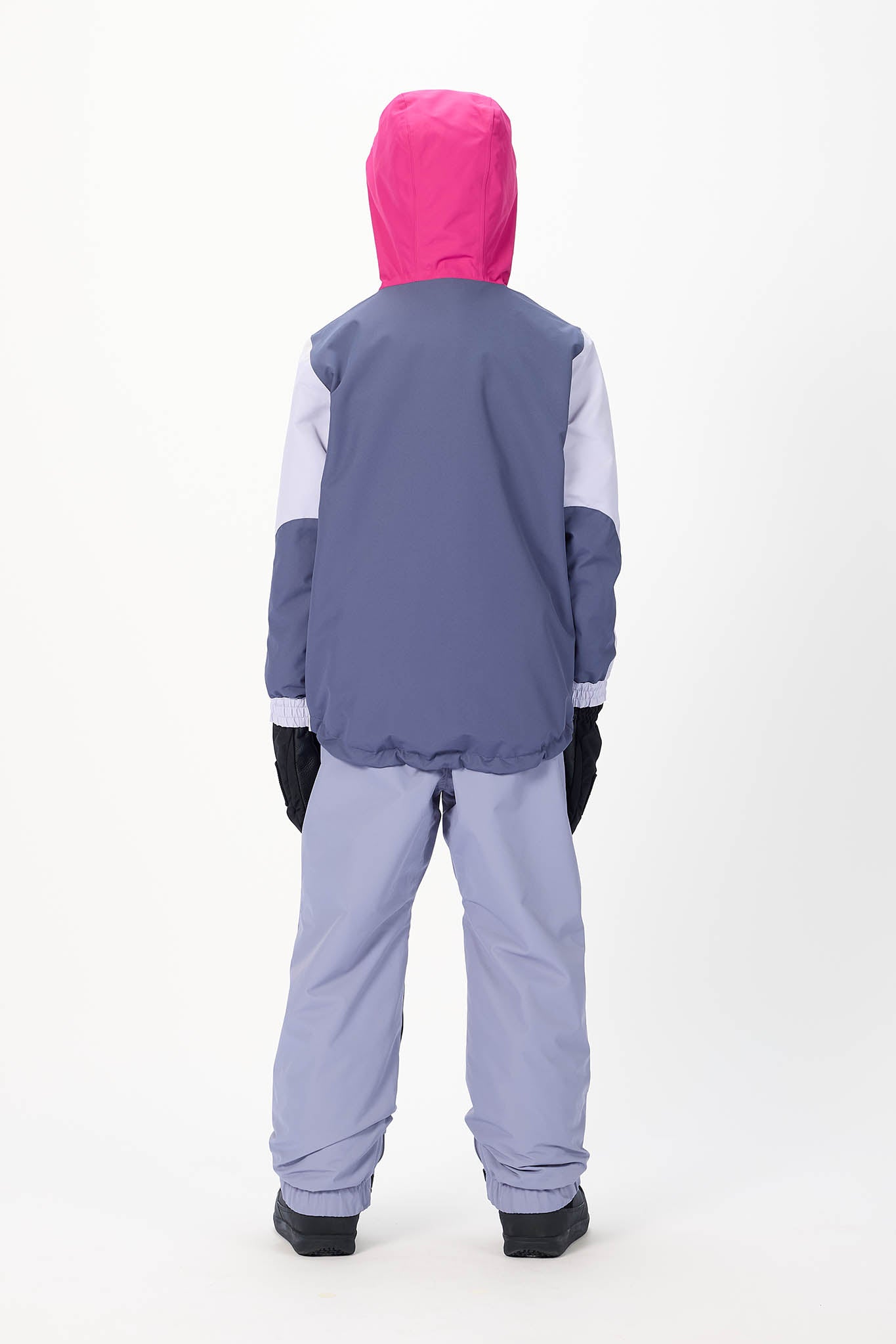 A child viewed from the back wearing a color-blocked hooded jacket with a bright pink hood, slate blue body, and white sleeves, paired with light lavender pants and black gloves, against a plain white studio background.