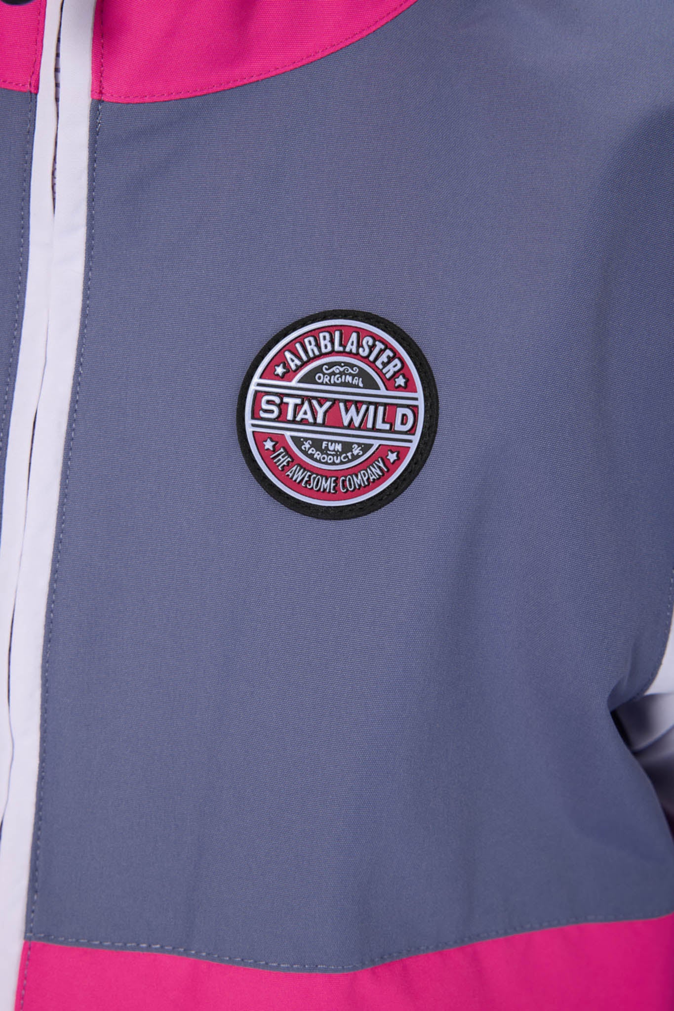 Close-up of a colorblock jacket in slate blue, hot pink, and white, featuring a circular 'Fairblaster Stay Wild' patch on the chest.