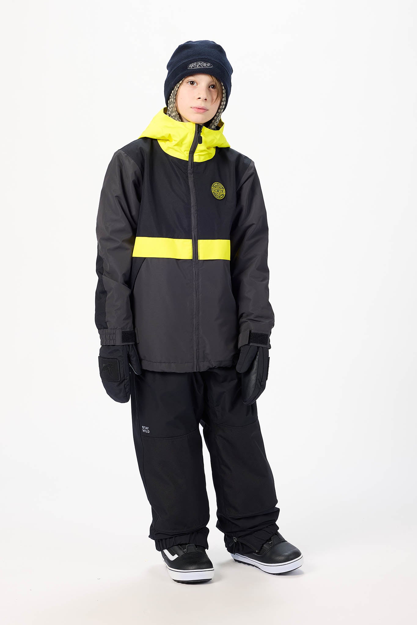 A child wearing a black ski jacket with a bright yellow collar and horizontal stripe, black snow pants, black gloves, a black beanie, and black sneakers, standing against a plain white studio background.