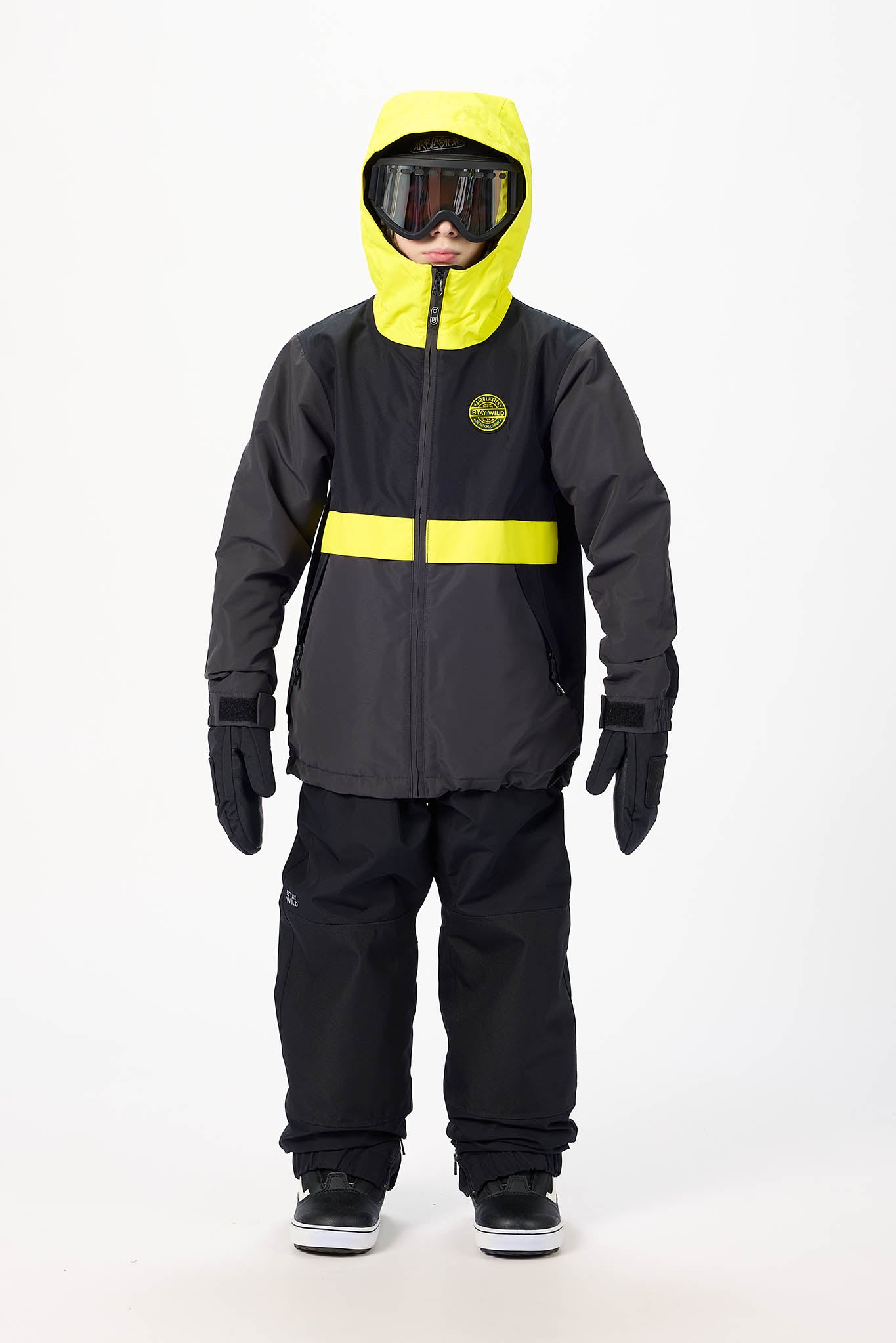 A child wearing a black ski jacket with a bright yellow hood and horizontal reflective stripe, paired with black snow pants, gloves, and goggles, standing against a white studio background.