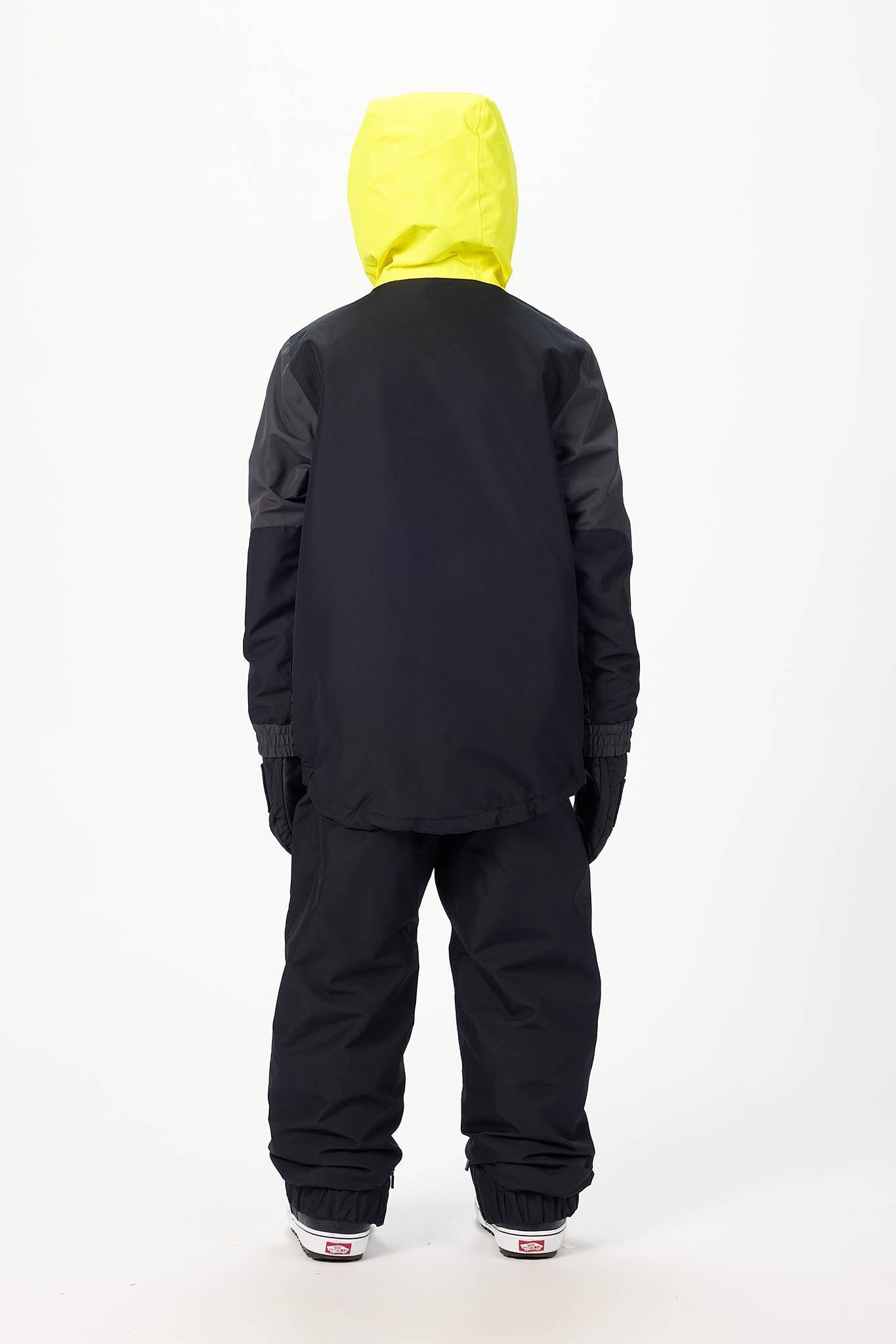 A child viewed from the back wearing a black snow jacket with a bright neon yellow hood and matching black snow pants, standing against a plain white studio background.