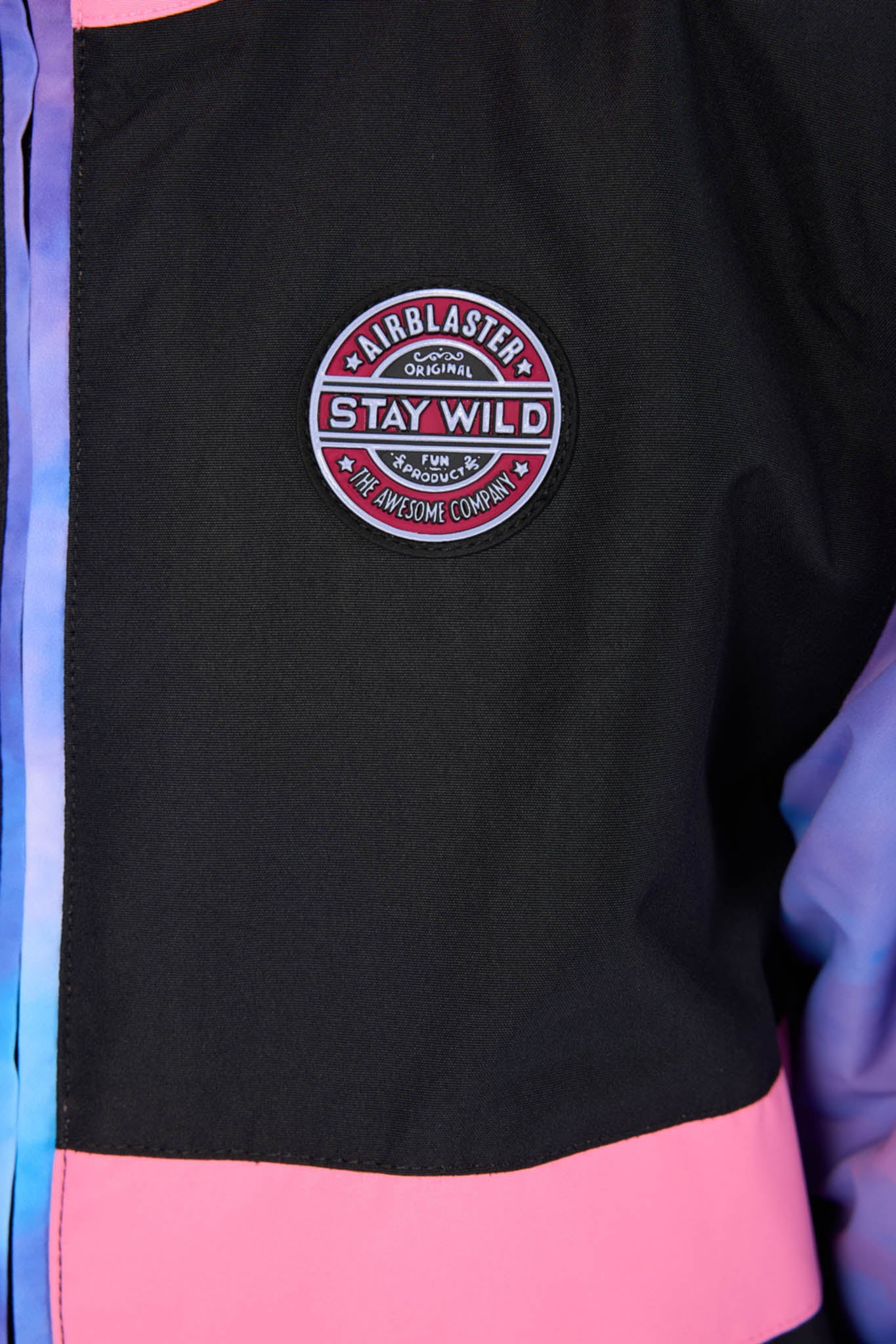 Close-up of a black jacket with a pink horizontal band at the bottom and a circular embroidered patch reading 'Airblaster Stay Wild' on the chest.