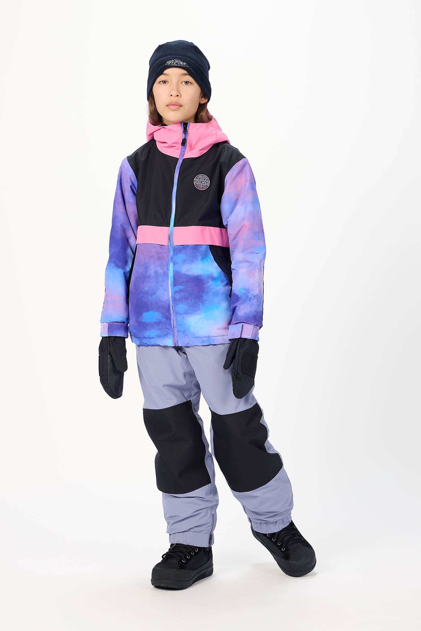 A child model wearing a colorful winter sports jacket with a purple, blue, and pink galaxy print, paired with gray and black snow pants, black gloves, and a black beanie, standing against a plain white studio background.