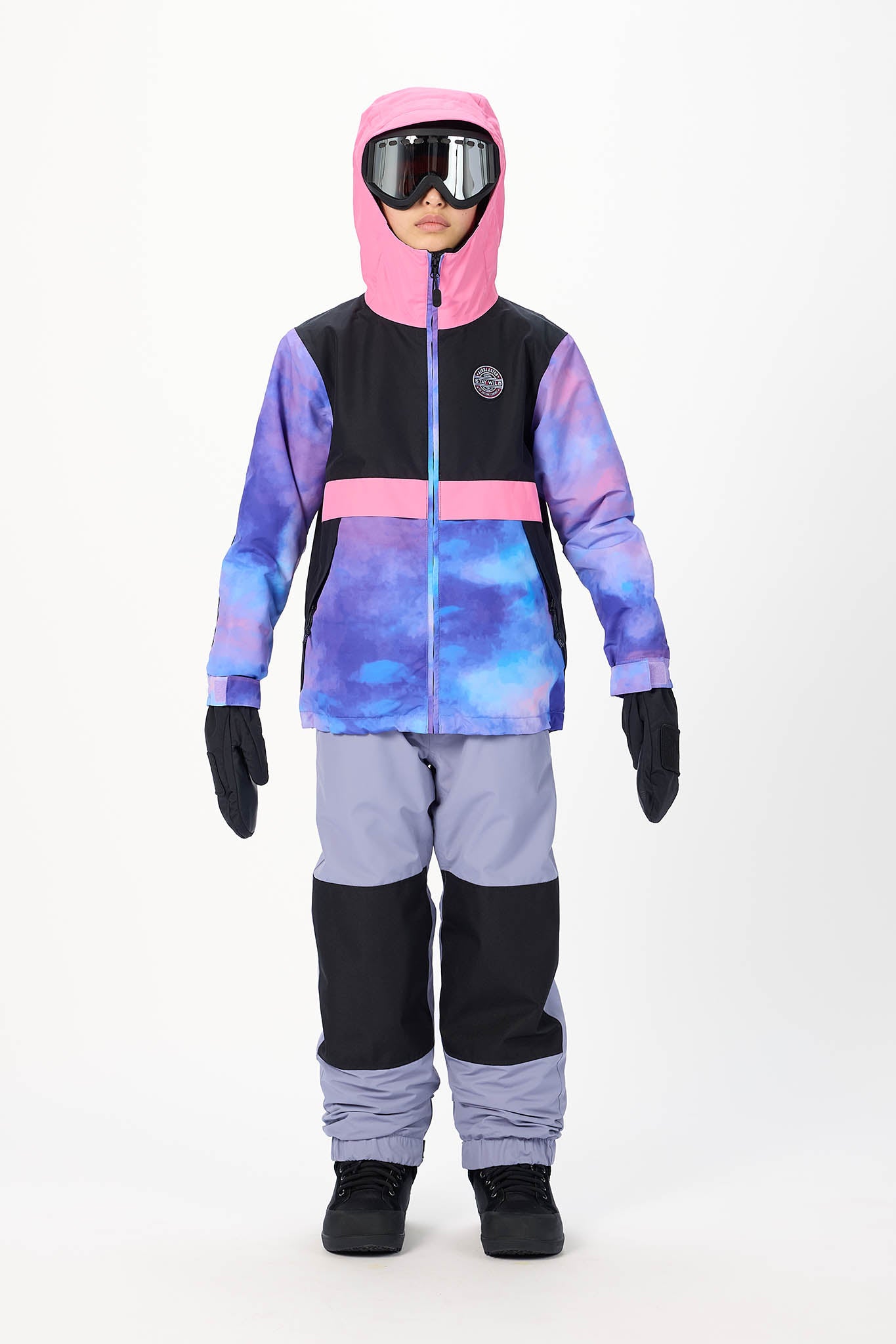 A child wearing a hooded ski jacket with a pink hood, black and blue-purple watercolor pattern body, pink chest stripe, lavender pants, black gloves, and goggles against a white studio background.