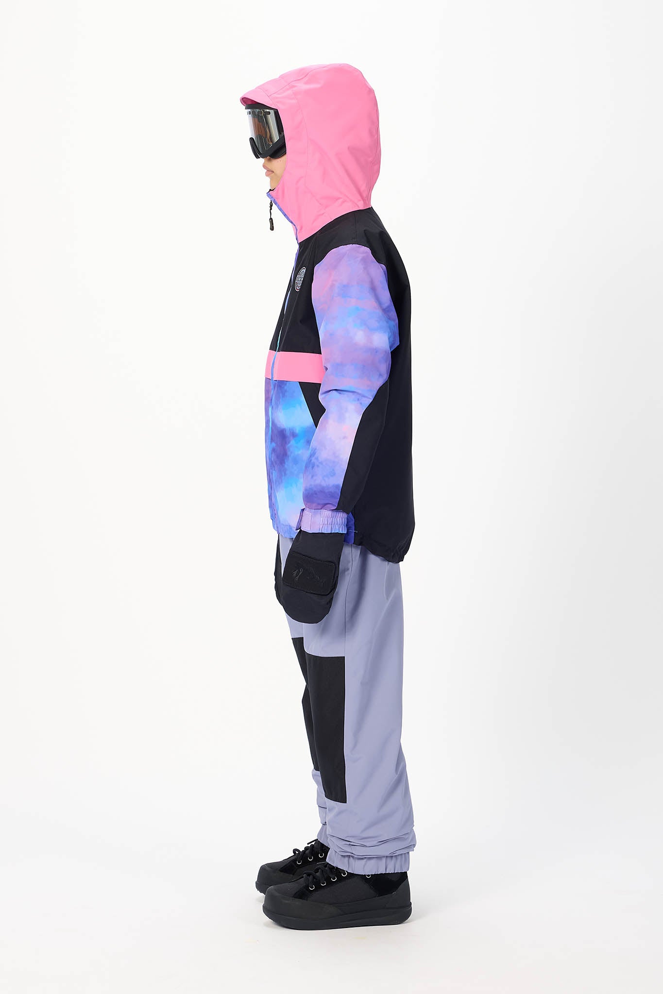 A child model shown in profile wearing a color-blocked ski jacket with a pink hood, black upper section, and blue/purple galaxy-patterned lower body, paired with light gray pants and black ski boots, against a plain white studio background.